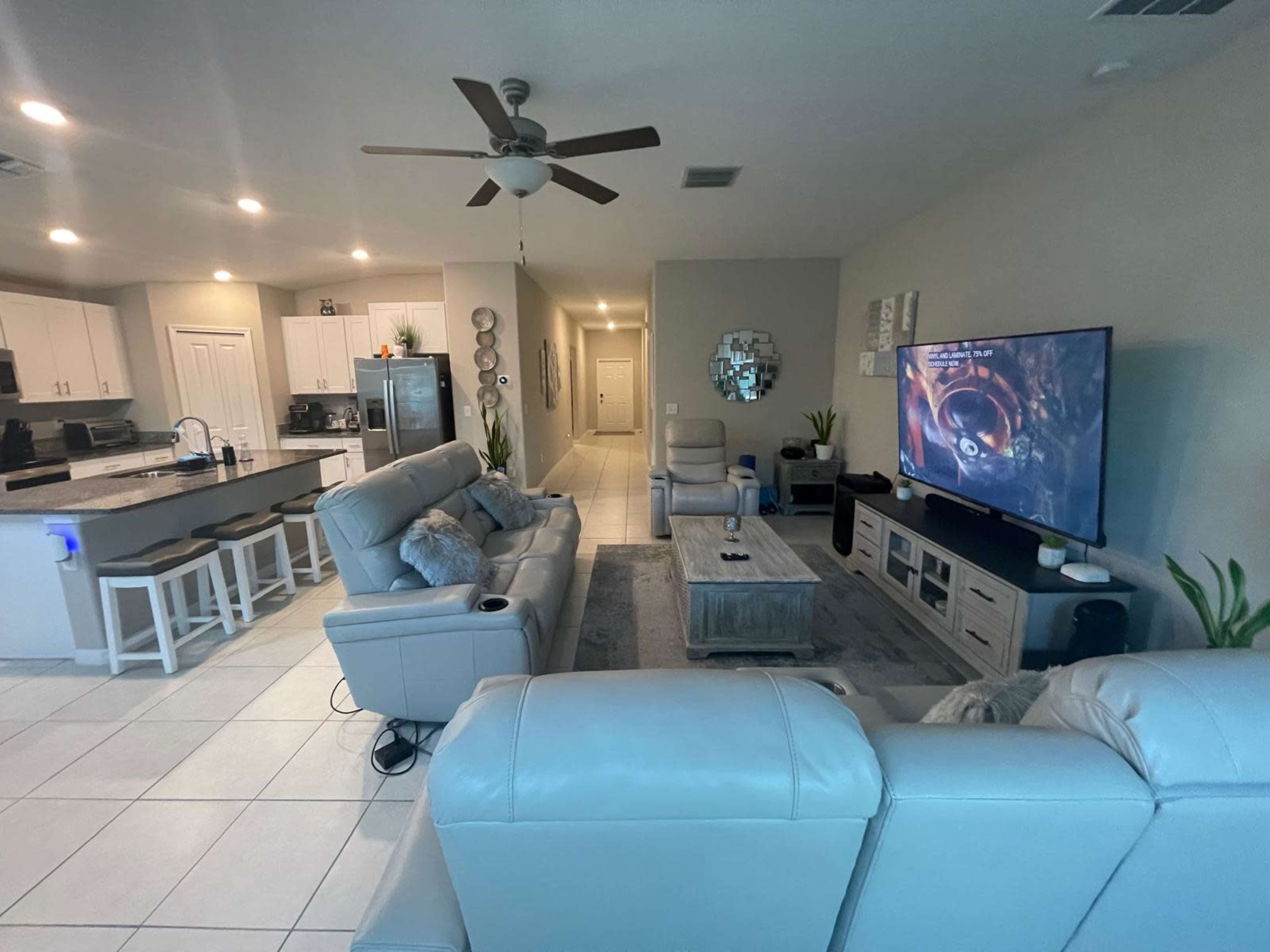The image shows a modern living room with a light-colored sofa, a large TV on a stand, and an open kitchen area in the background.