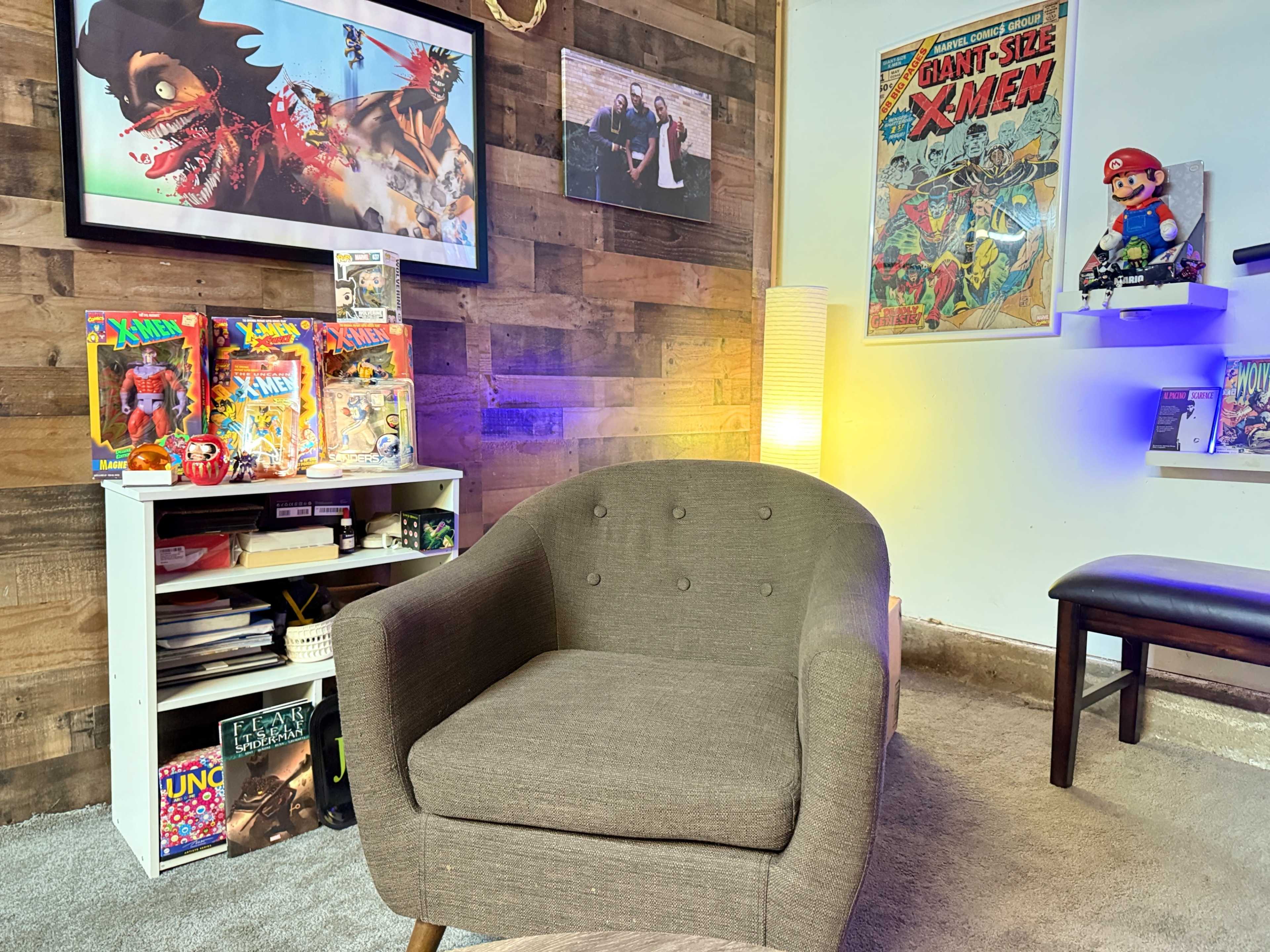 The image shows a cozy room featuring a gray armchair, a variety of colorful comic books and collectibles, and framed artwork on the walls.