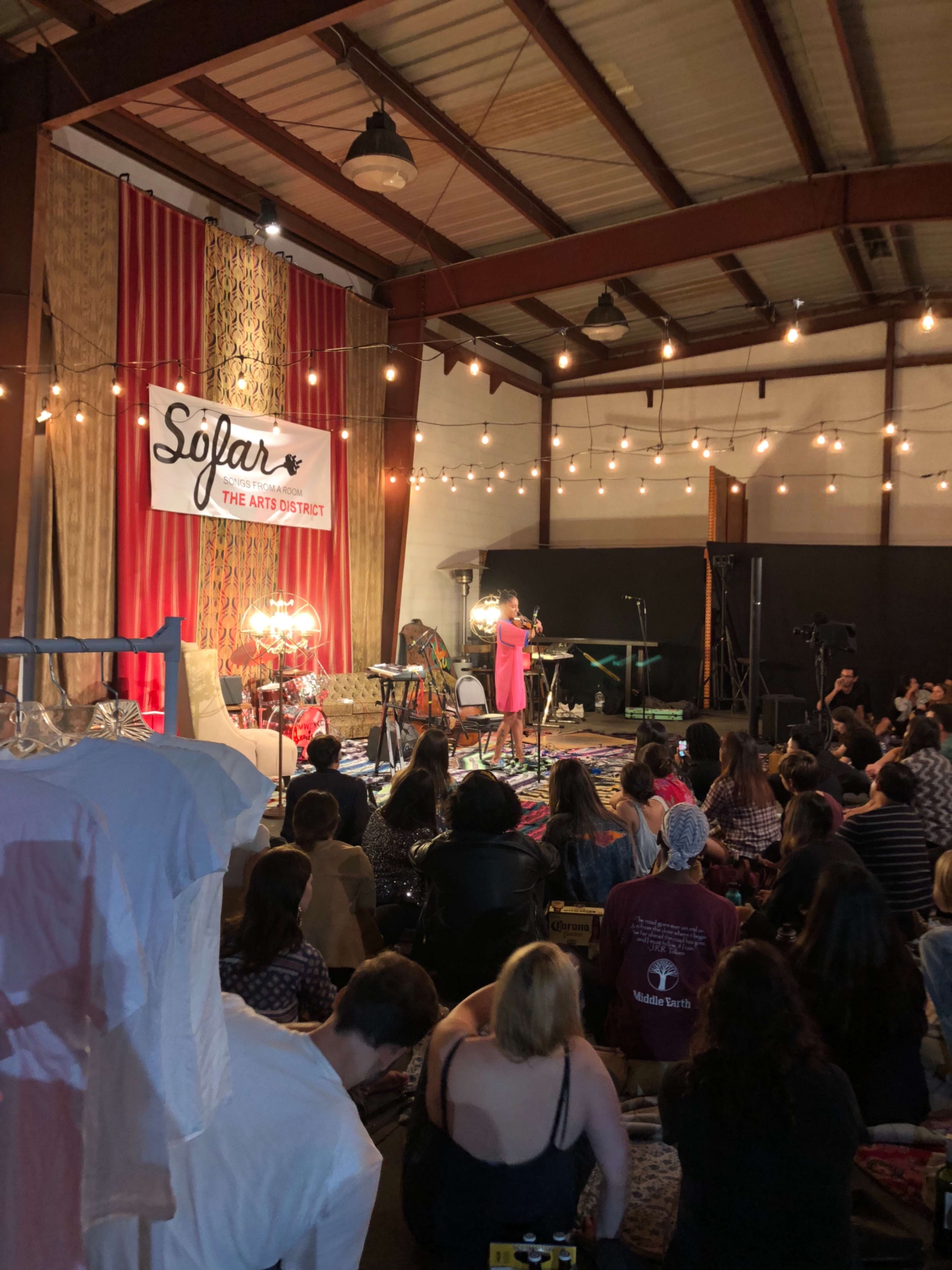 A performer stands on stage in front of an audience in a warmly lit indoor space decorated with string lights and eclectic furnishings.