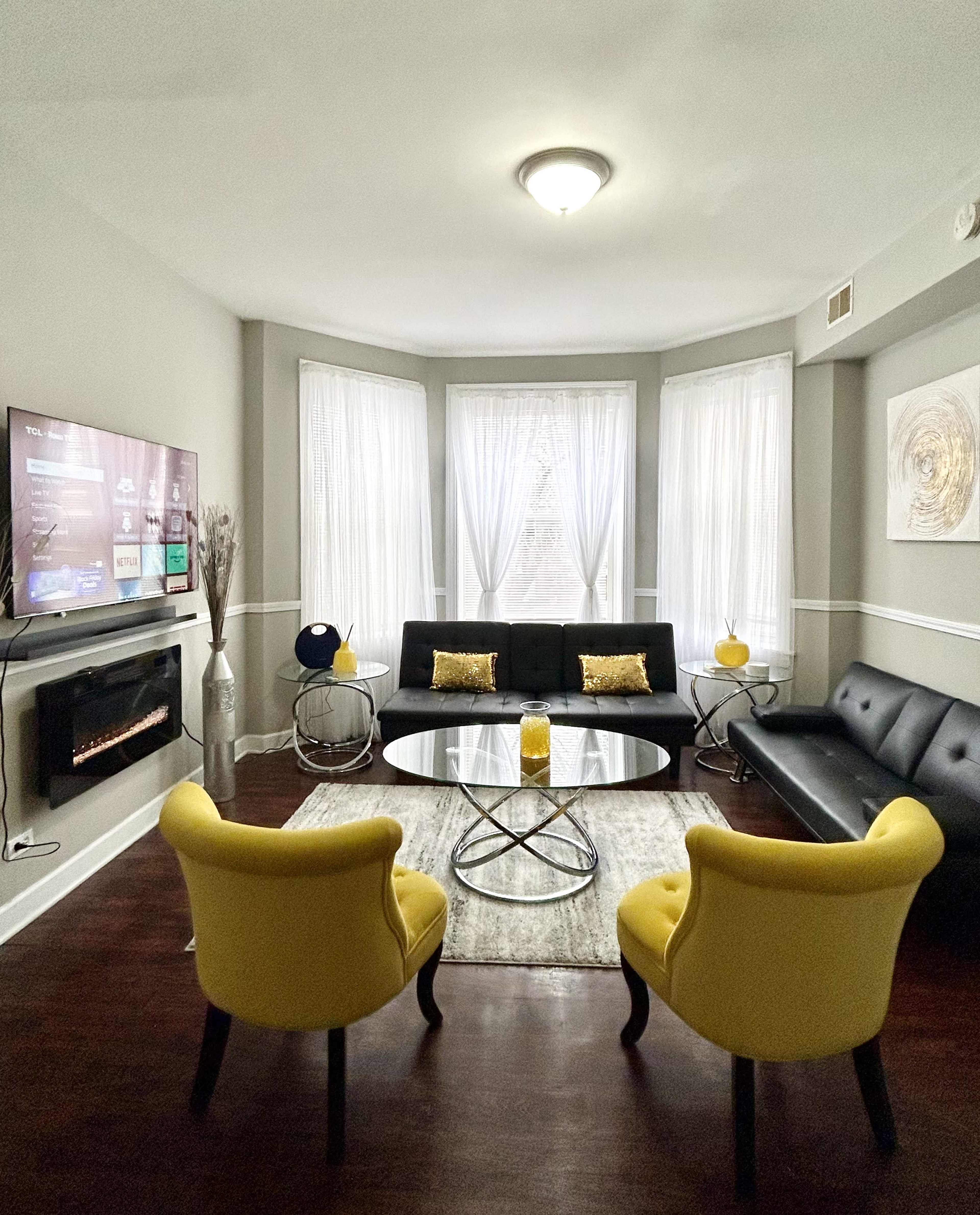 The image shows a modern living room featuring a black sofa, a glass coffee table, two yellow chairs, and a television mounted on the wall, with large windows draped in sheer curtains.