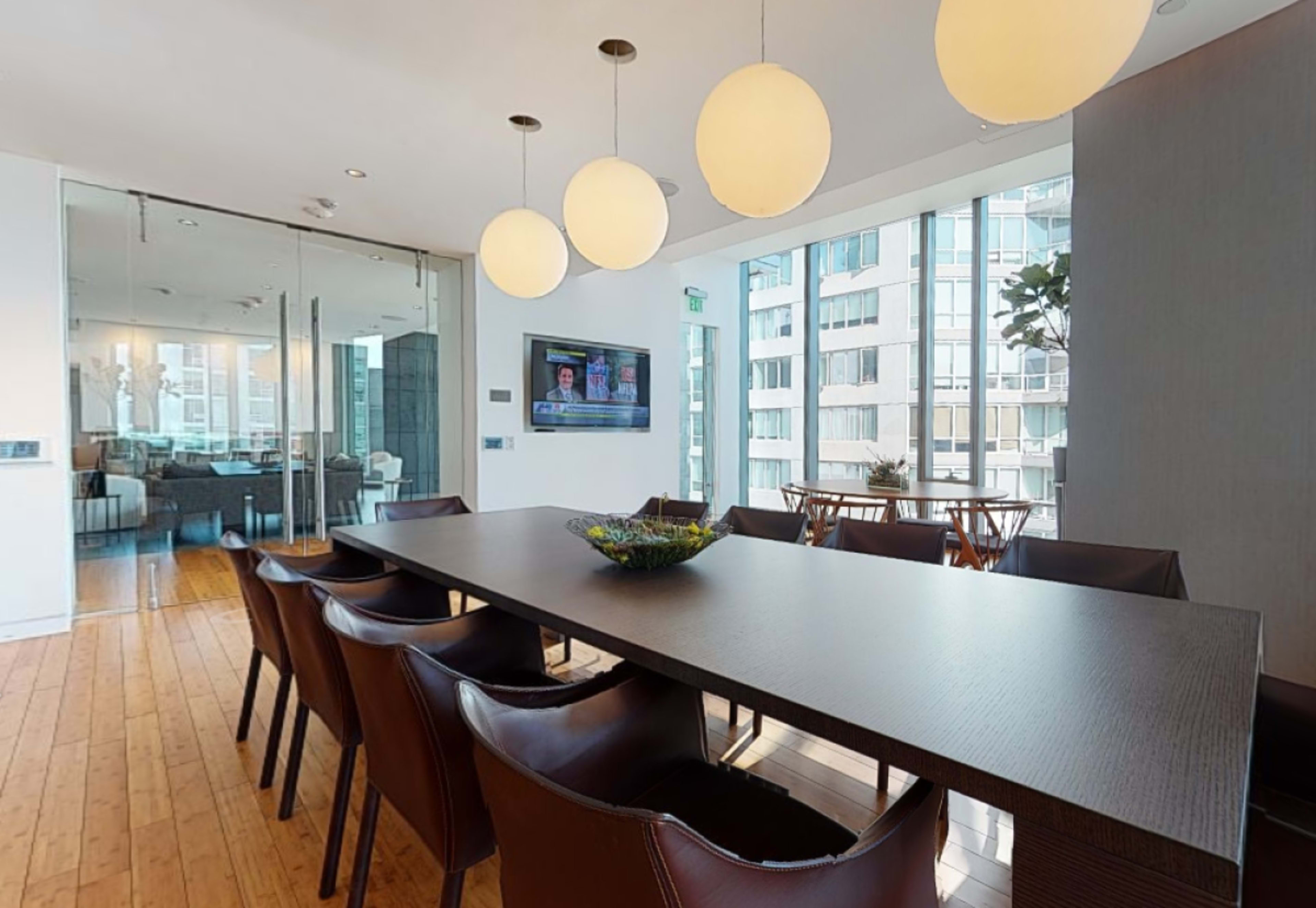 A modern dining area features a long wooden table with dark chairs, illuminated by hanging round lights and large windows overlooking a cityscape.