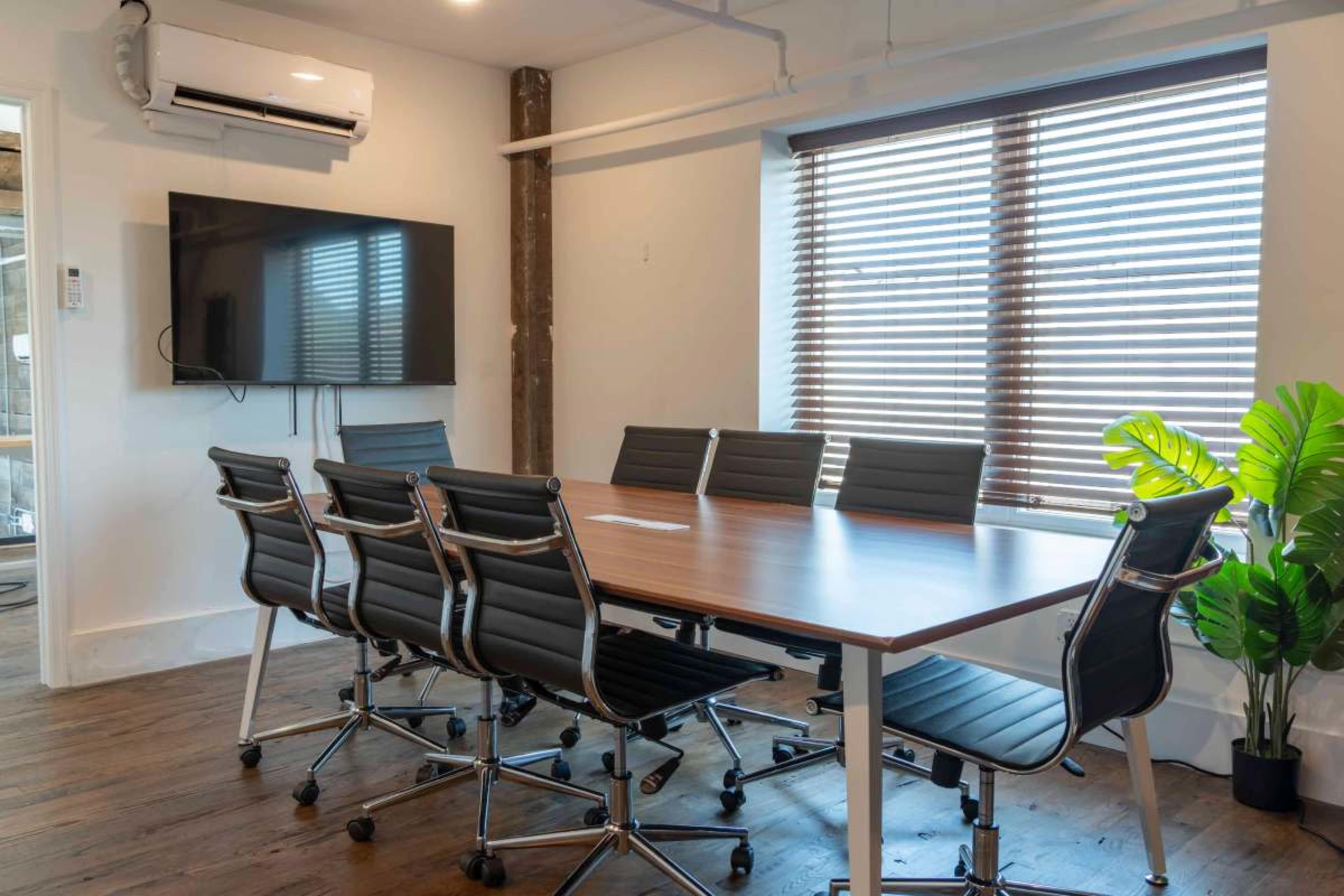 A modern conference room features a large wooden table surrounded by black rolling chairs, with a wall-mounted TV and a window partially covered by blinds.
