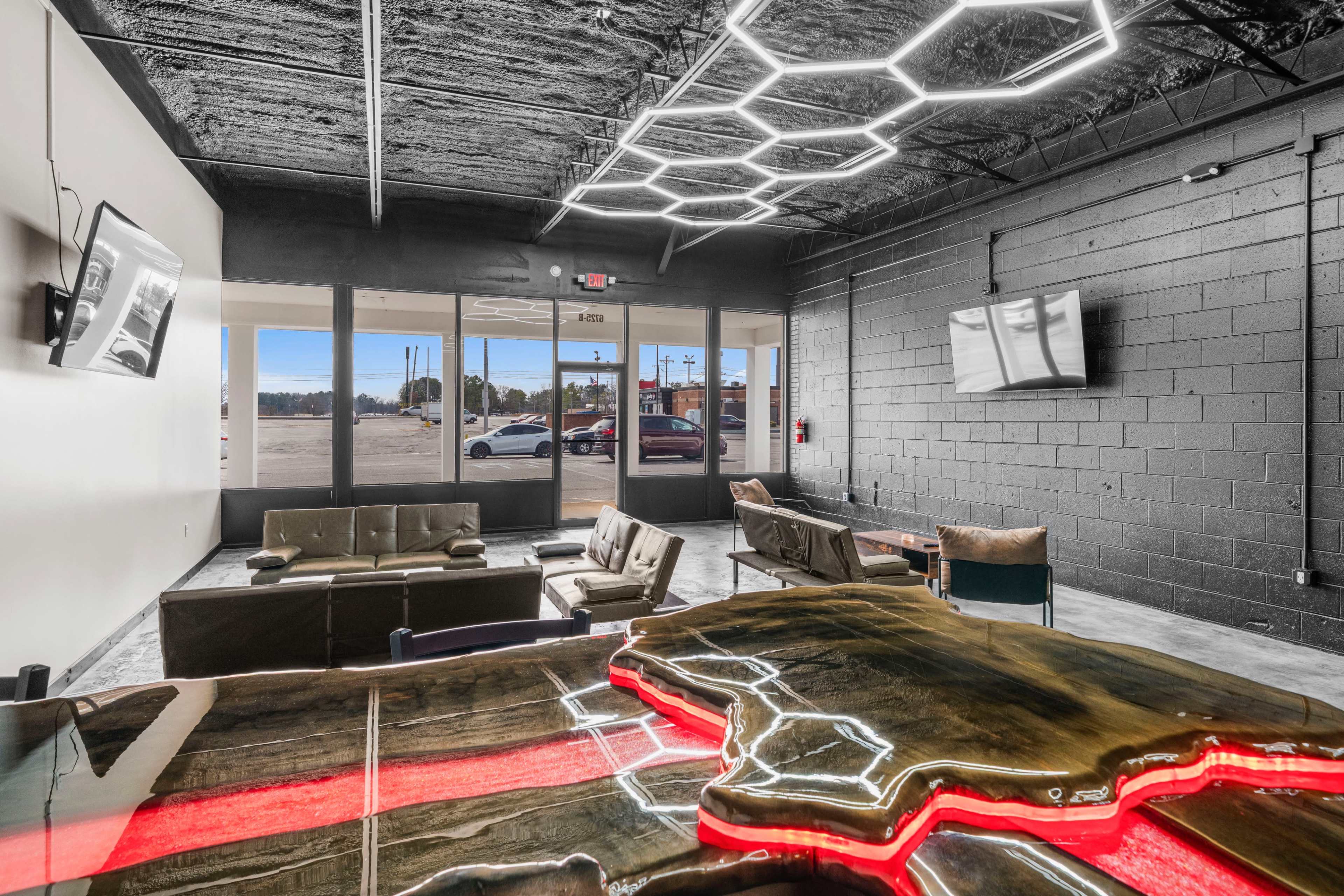 The image shows a modern lounge area with a unique wooden table featuring neon lighting, leather seating, and large glass doors leading to the outside.