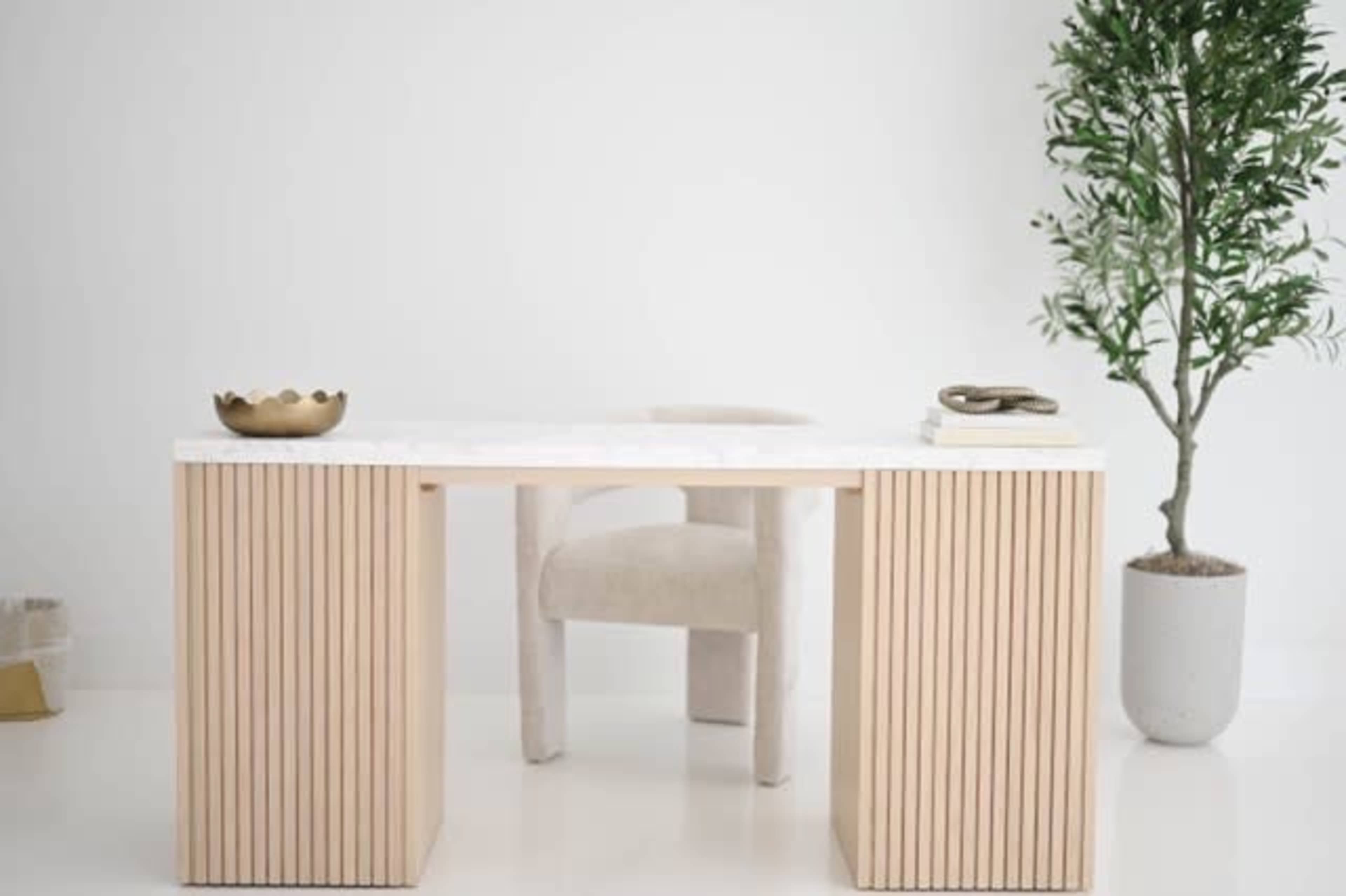 A minimalistic desk with vertical wood slats is positioned next to a plant and a chair in a bright, neutral-colored room.