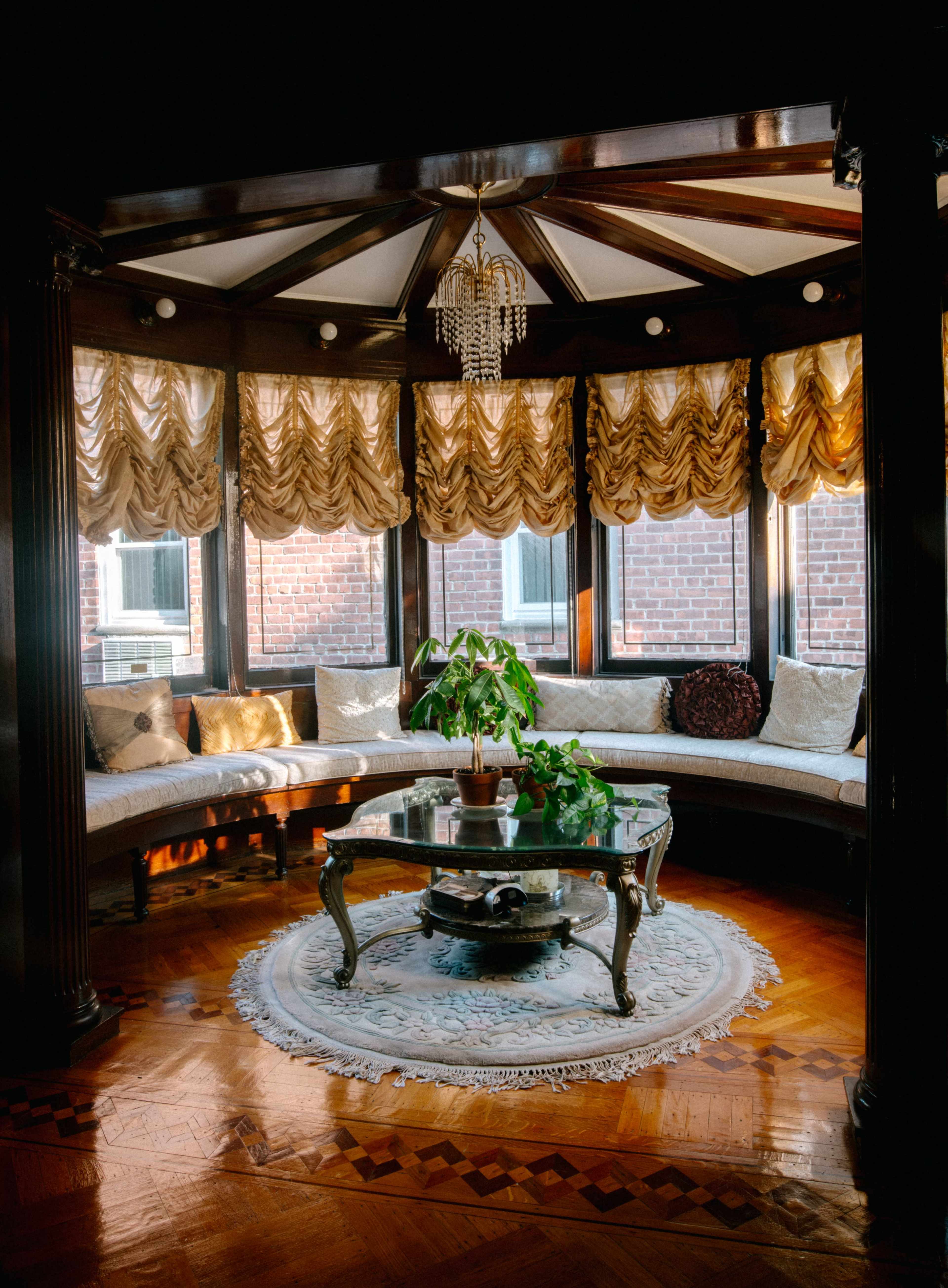 The image shows a sunlit circular sitting area adorned with plush seating, a glass coffee table, and a potted plant, featuring elegant drapery and wooden architecture.