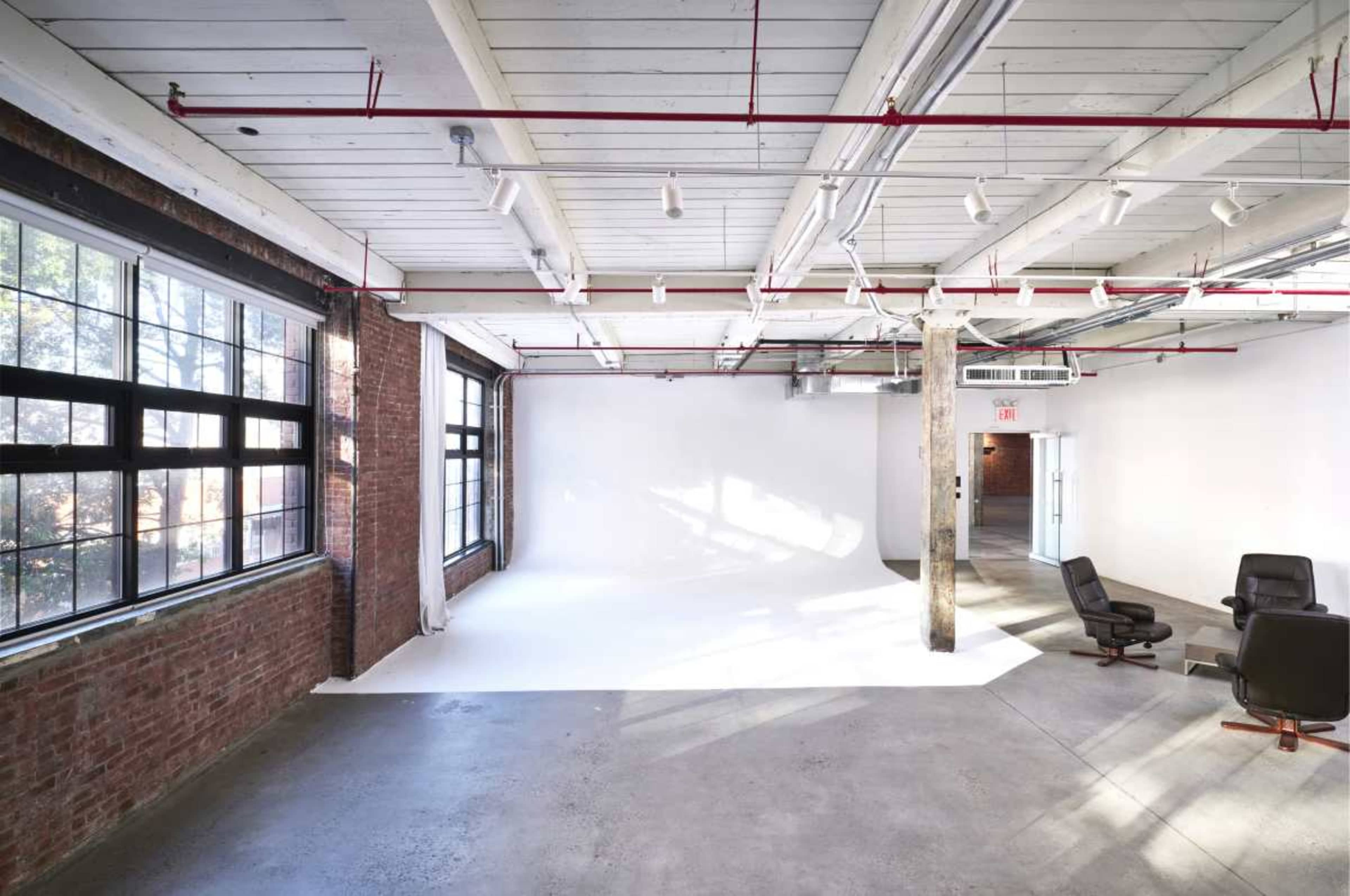 The image shows a spacious, industrial-style studio with large windows, exposed brick walls, and a white seamless backdrop.