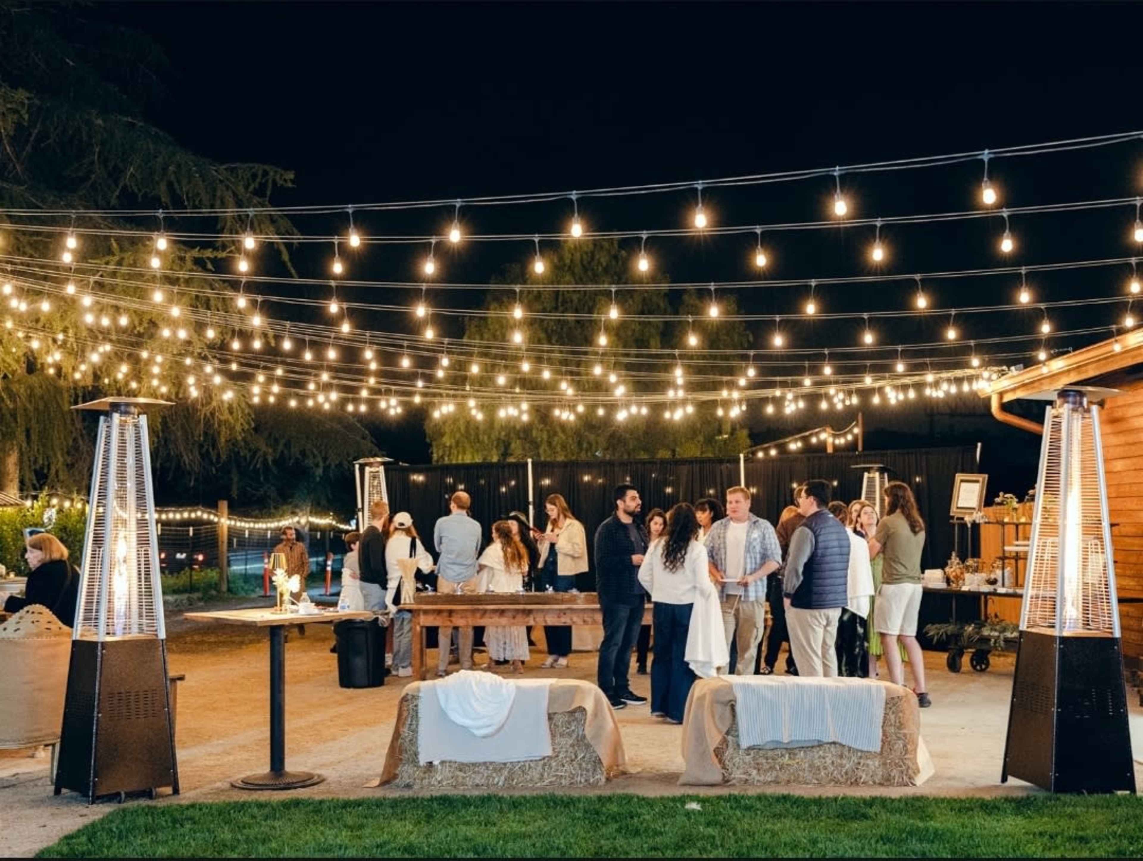 A group of people socializes under string lights at an outdoor event venue, with several outdoor heaters visible.