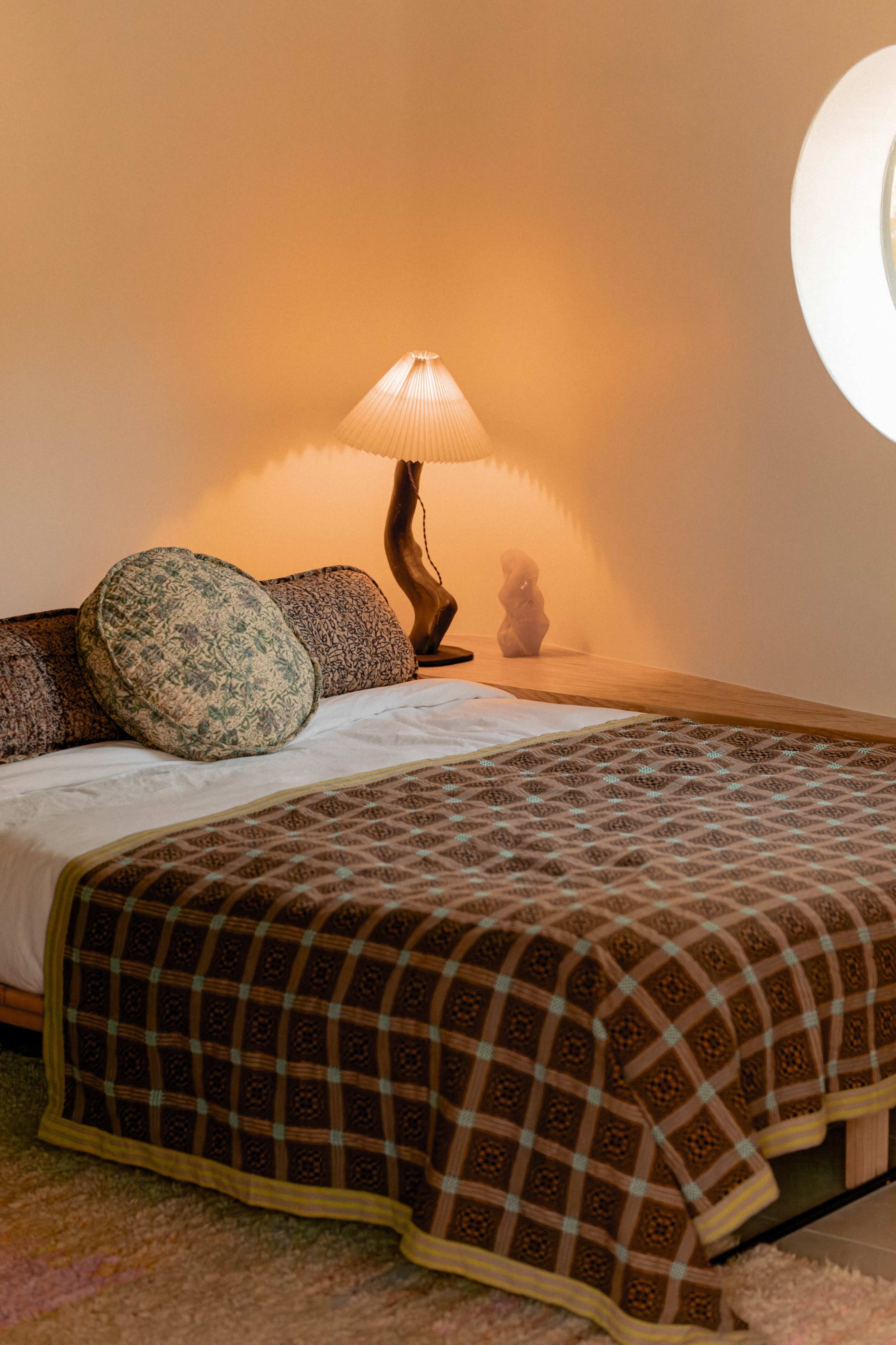 The image shows a bedroom with a bed covered in a patterned blanket, a decorative pillow, and a lamp casting a warm glow on the wall.