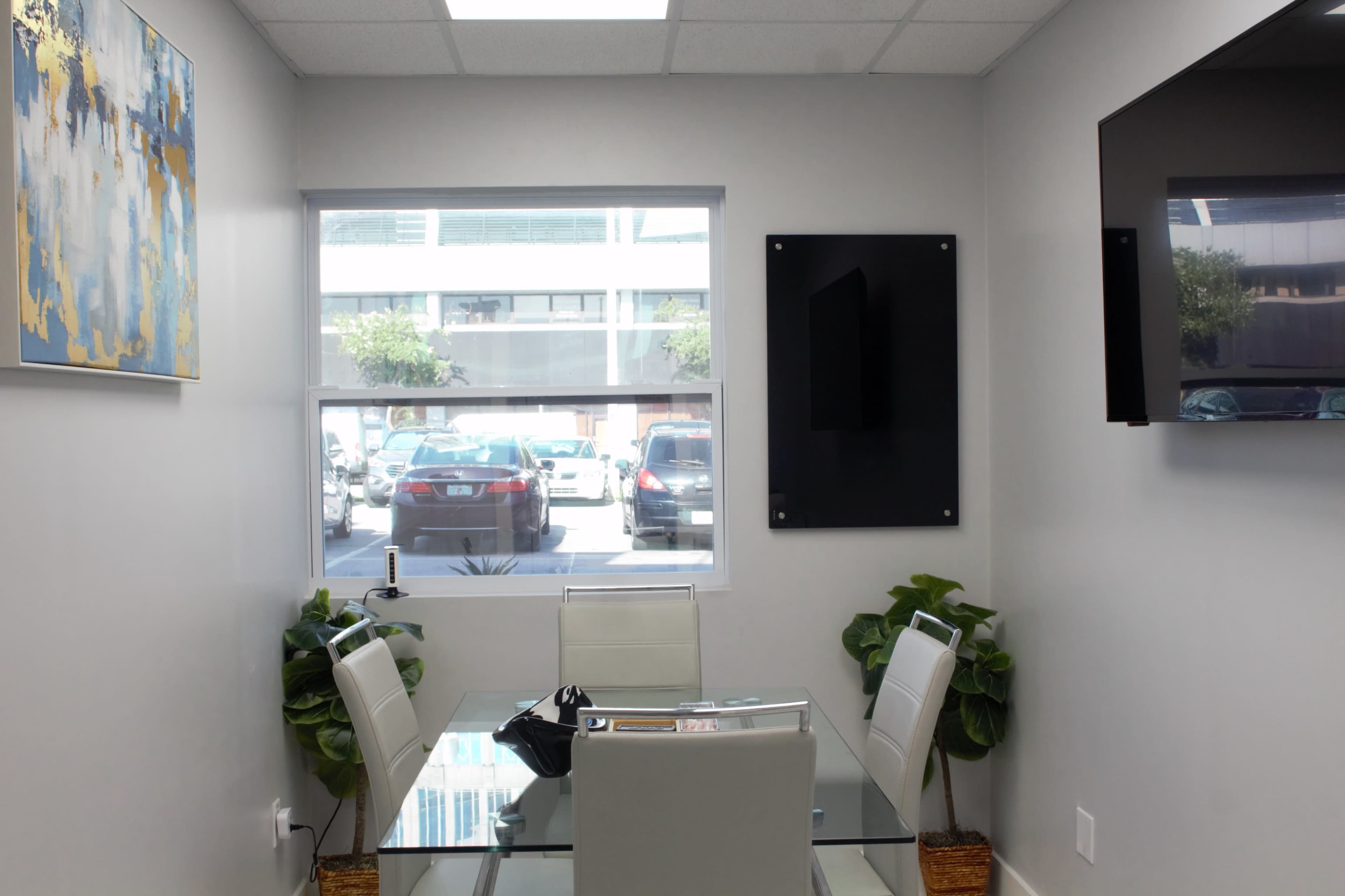 A modern conference room features a glass table surrounded by white chairs, with a large painting on the wall and a television mounted high, overlooking a view of parked cars outside.