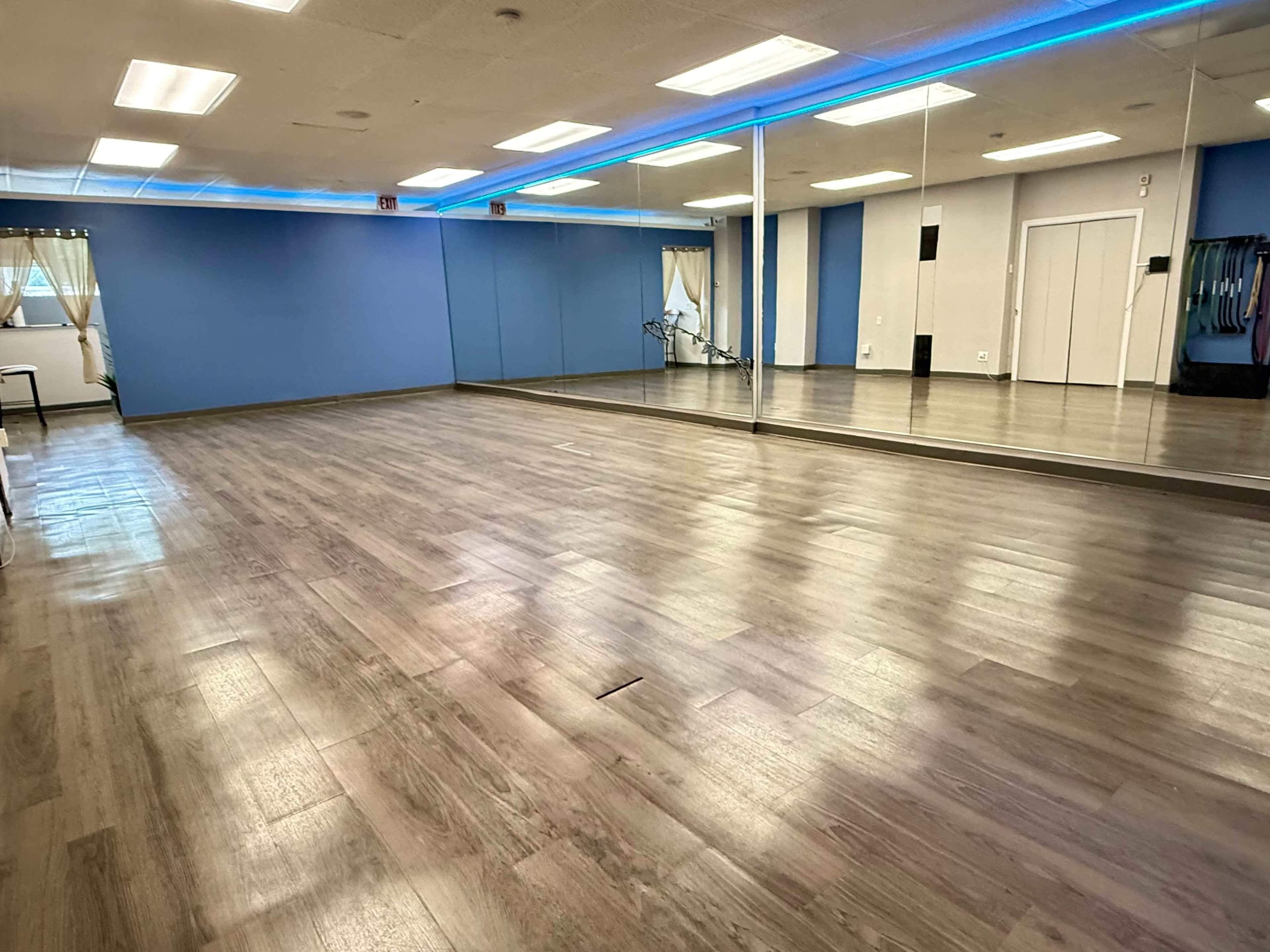 The image shows an empty fitness studio with blue walls, a large mirror, and wooden flooring.