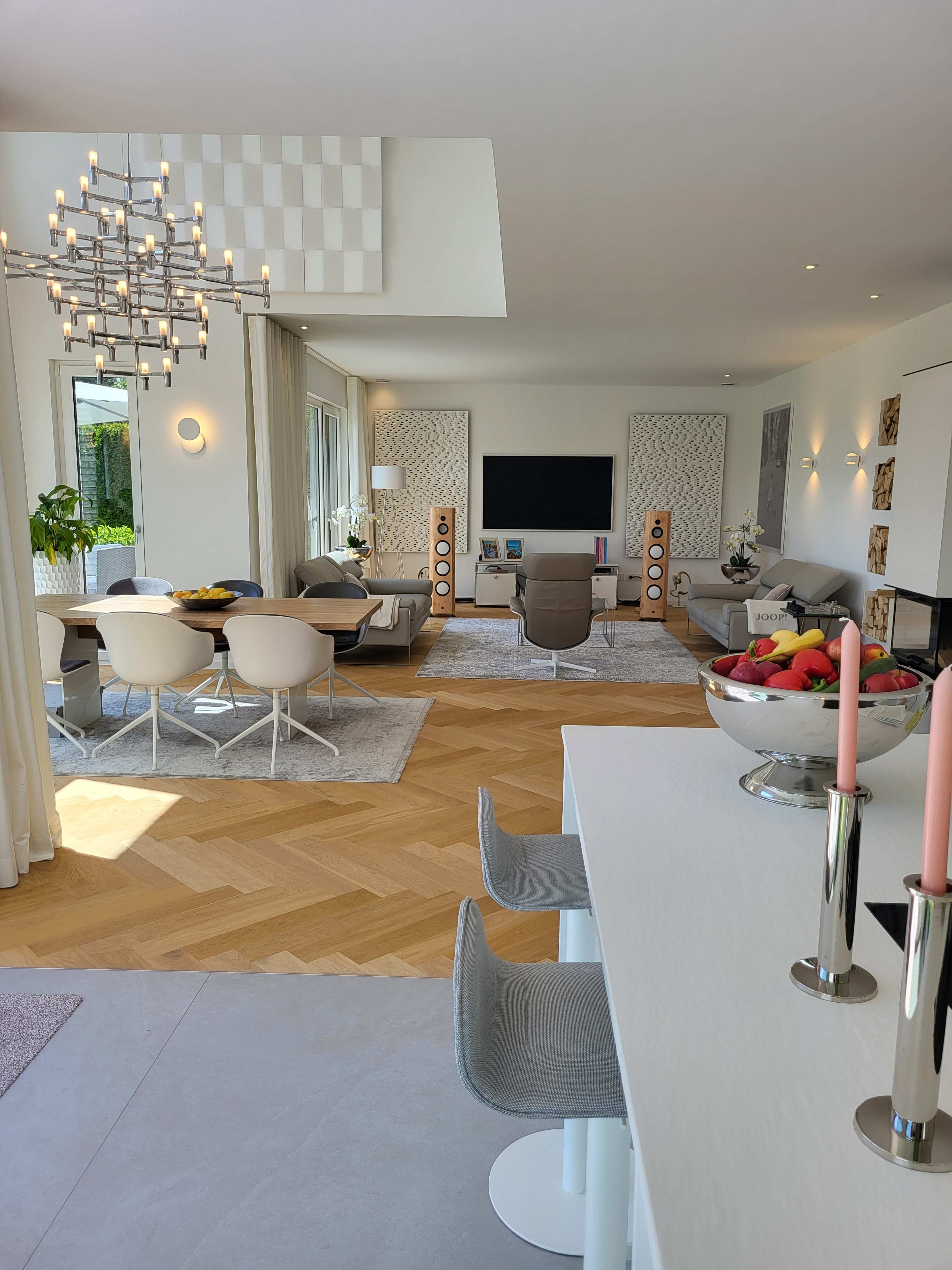 A spacious modern living room with a dining area, featuring large windows, a sleek entertainment setup, and contemporary furniture arranged around a light wood floor.