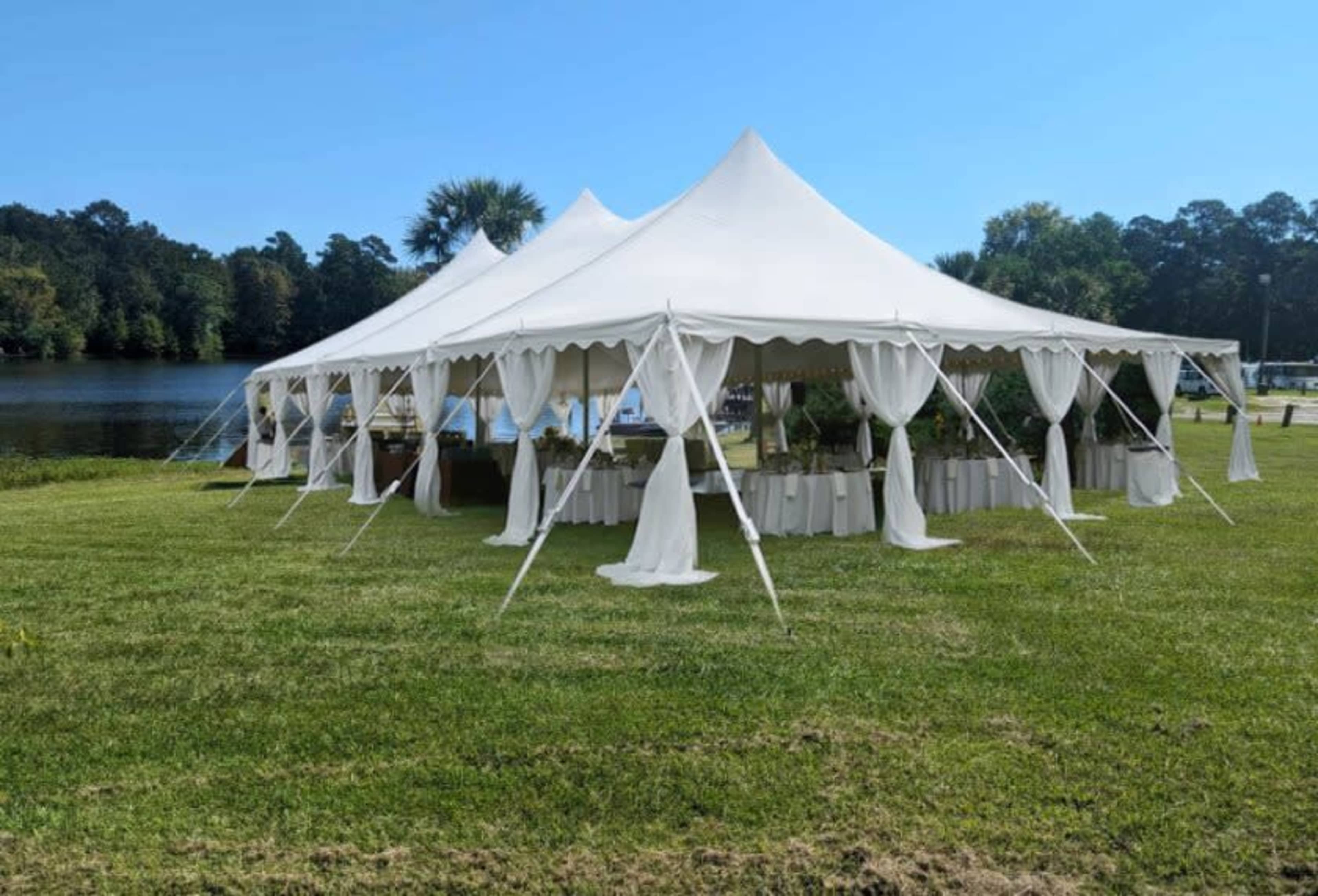 A large white tent with draped sides is set up on a grassy area beside a lake.