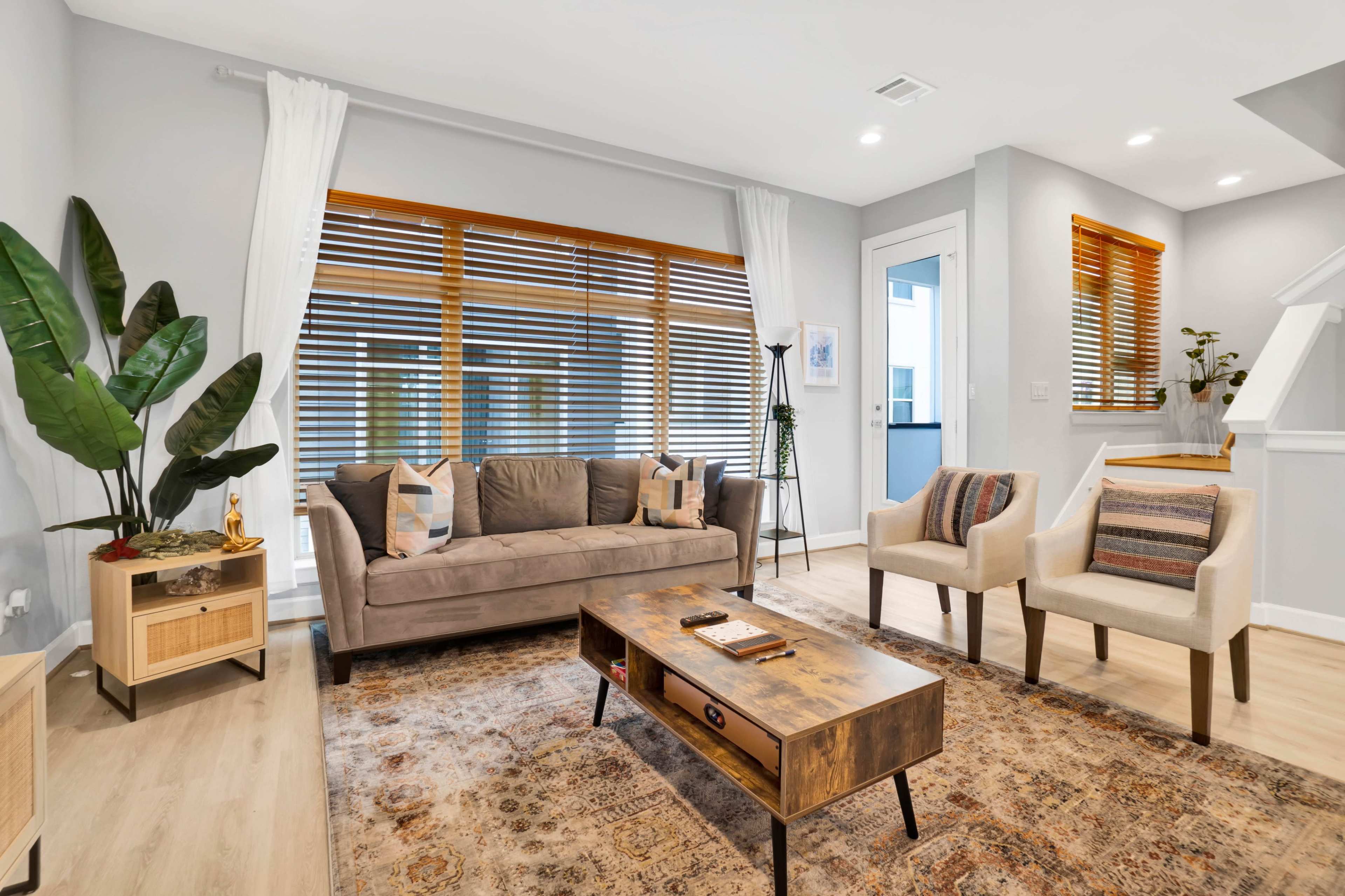 A modern living room features a sofa, two chairs, a coffee table, and large windows with wooden blinds.