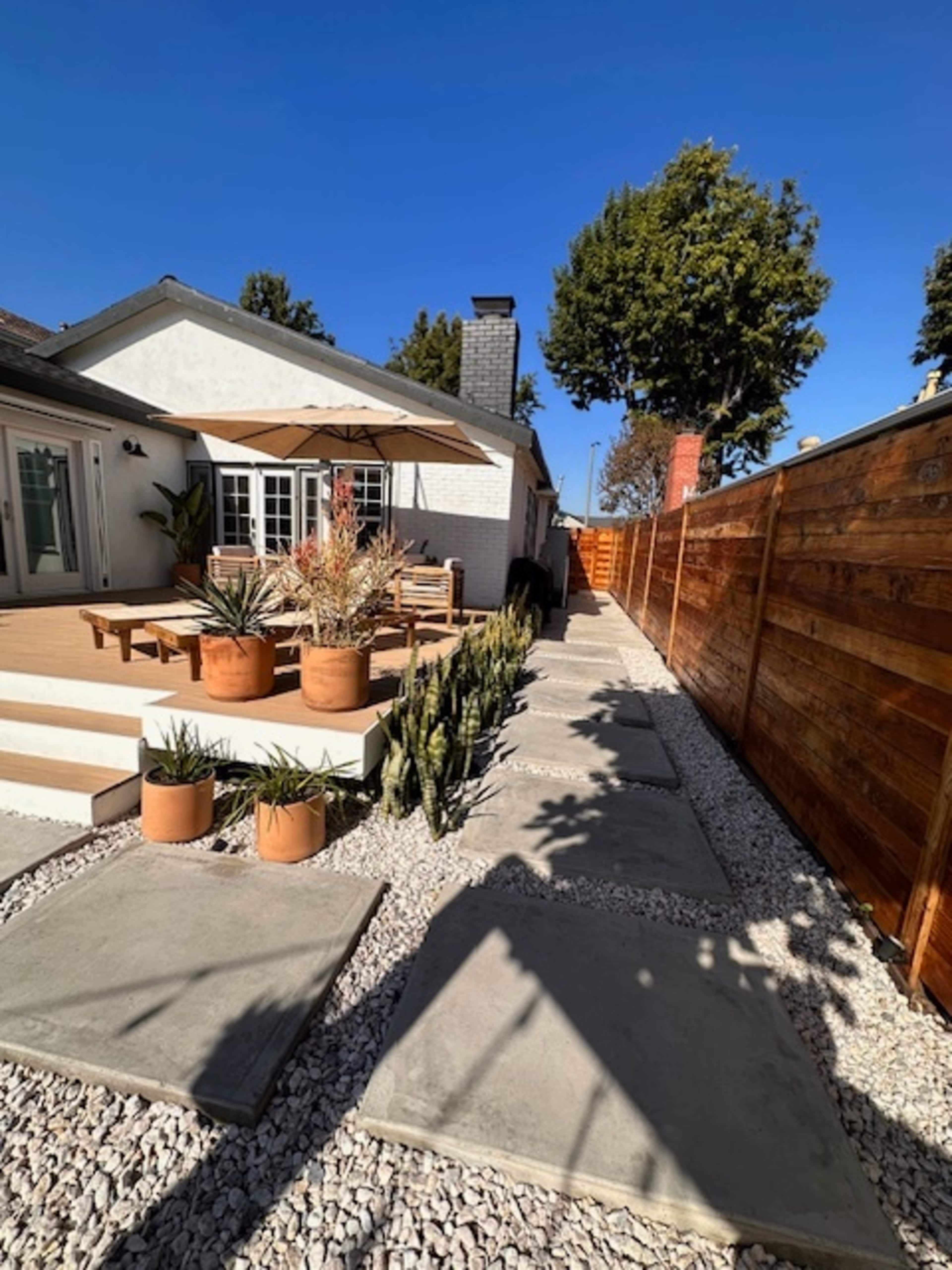 A pathway lined with concrete stepping stones and potted plants, leading to a home with a patio and a wooden fence.