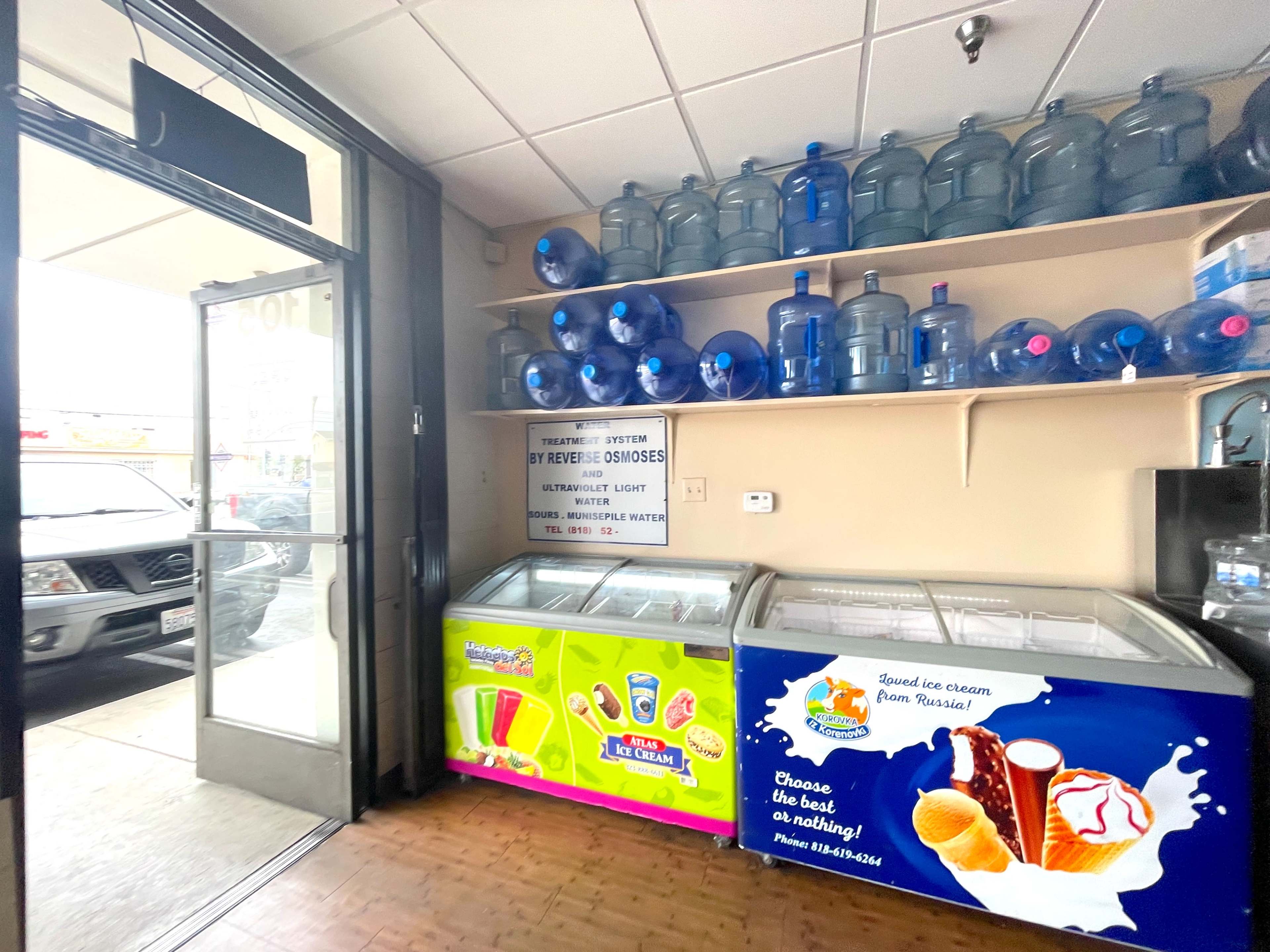 The image shows a corner of a store with two ice cream freezers and shelves of water bottles above them, alongside a glass door leading outside.