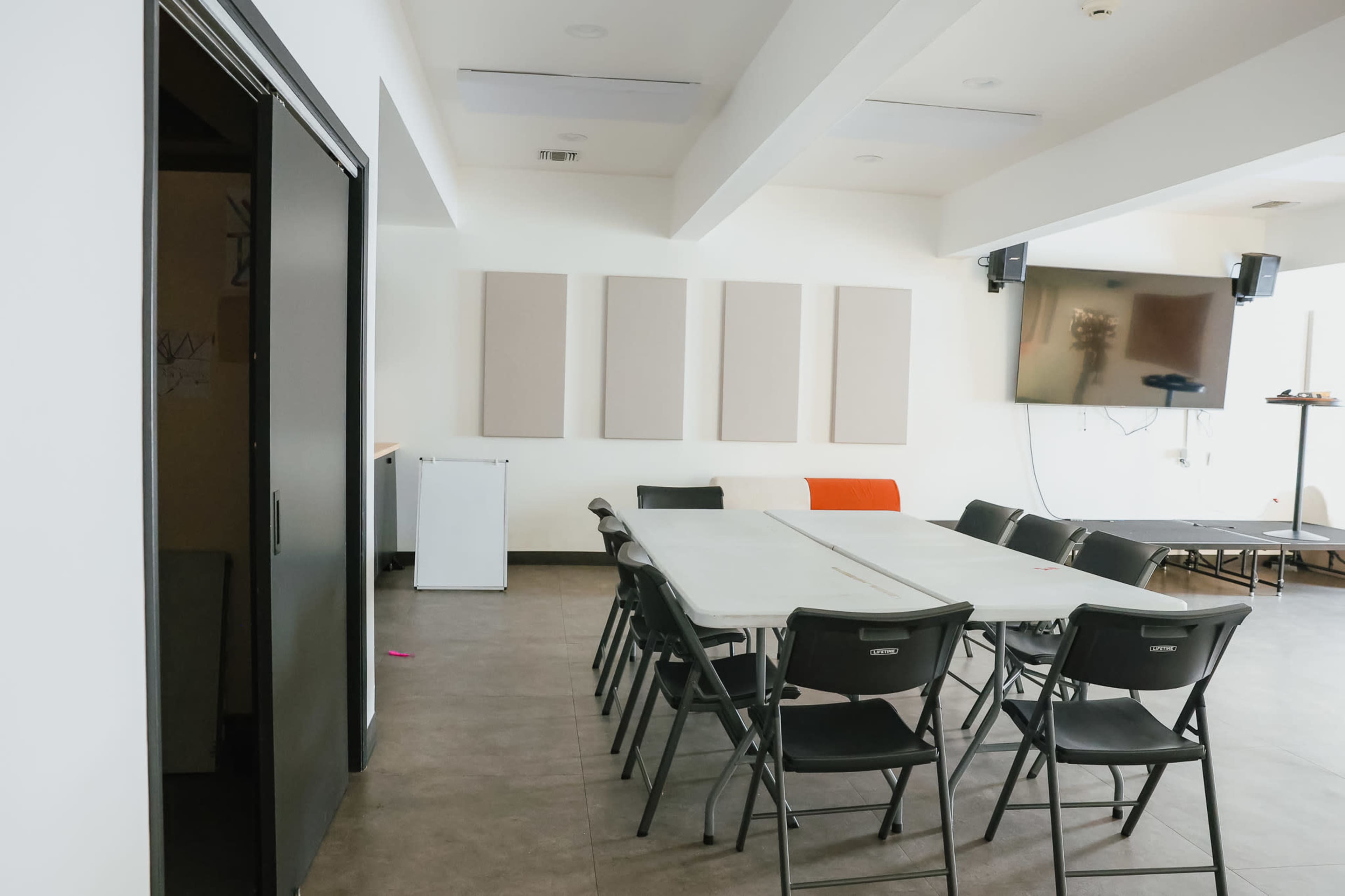 The image shows a spacious meeting room with a rectangular table surrounded by black folding chairs, a mounted TV, and soundproof panels on the walls.