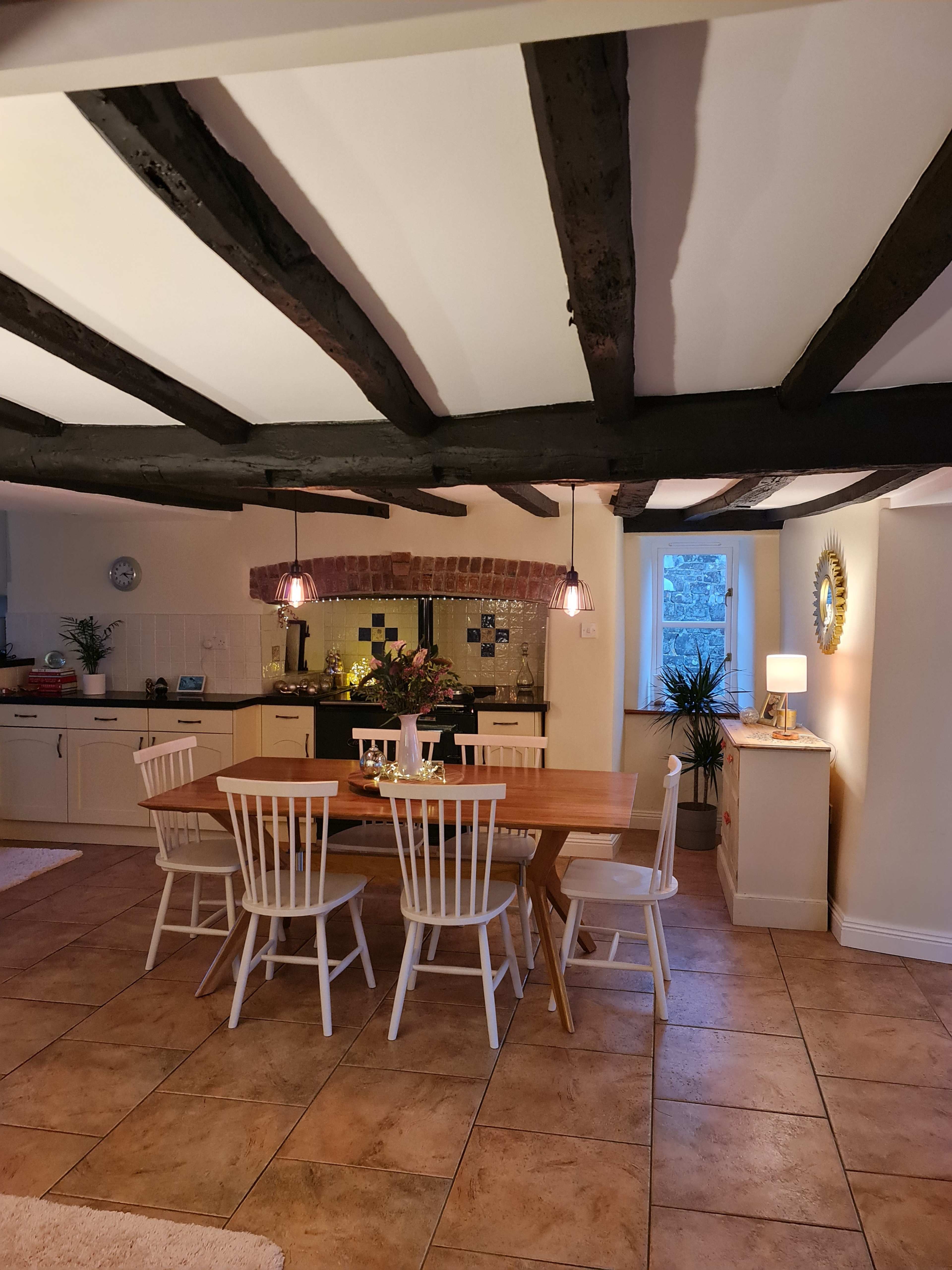 A dining area features a wooden table surrounded by six white chairs, with exposed beam ceilings and a kitchen visible in the background.
