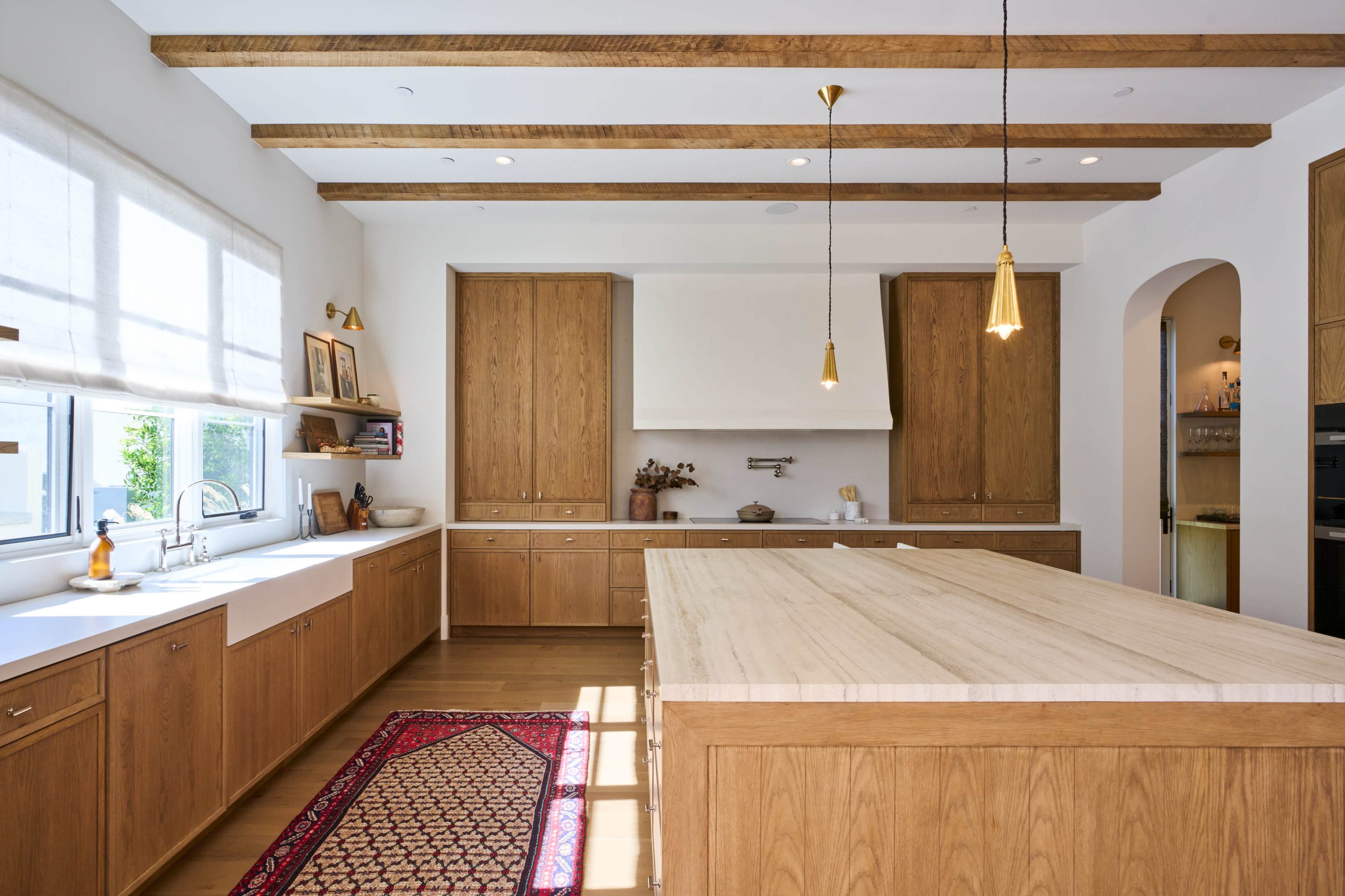 The kitchen features wooden cabinetry, a central island with a light countertop, and wooden beams along the ceiling, all arranged in a bright and spacious layout.