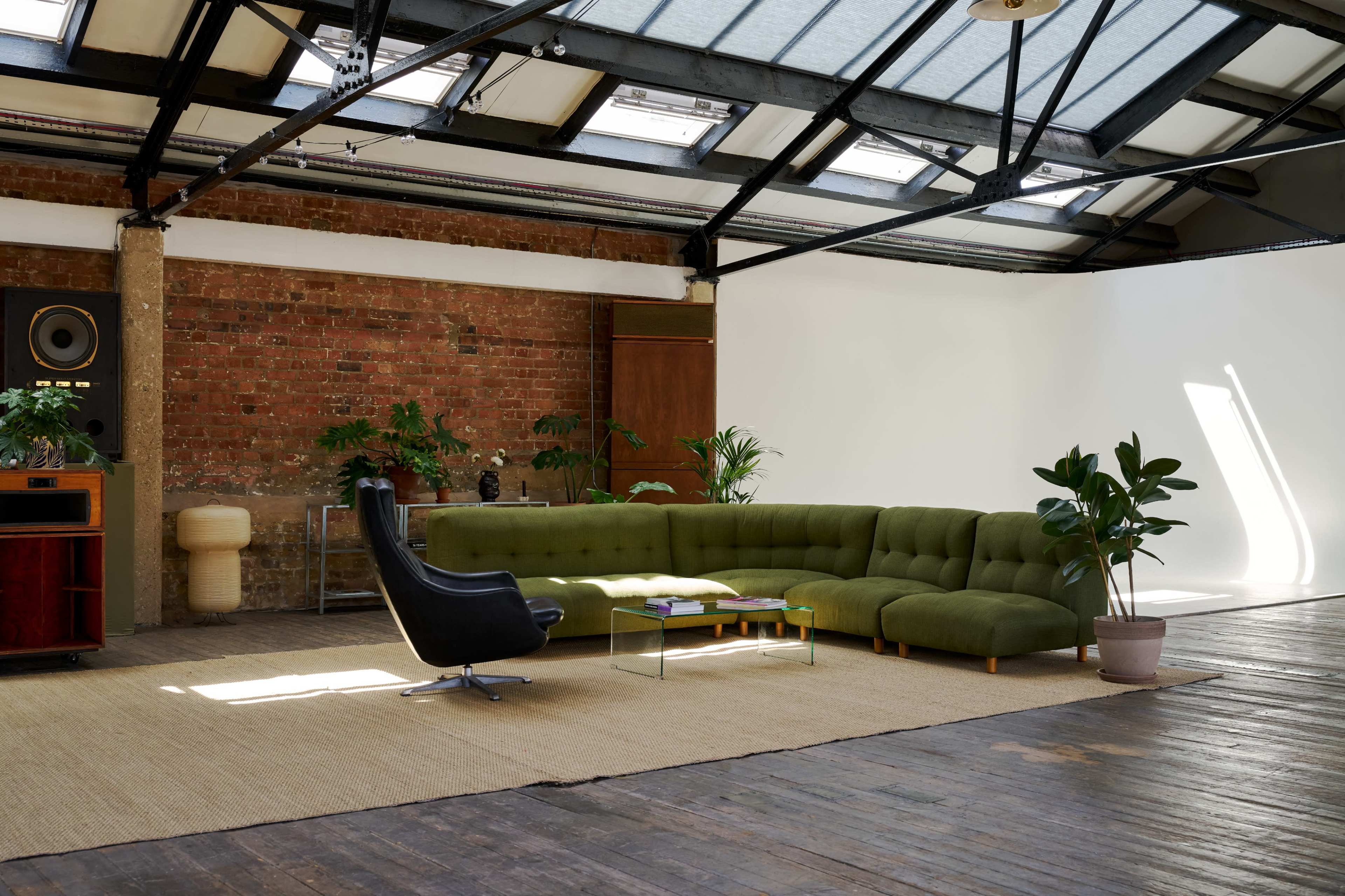 The image shows a spacious interior with a green sectional sofa, a black chair, and several plants against a backdrop of a brick wall and a white space.
