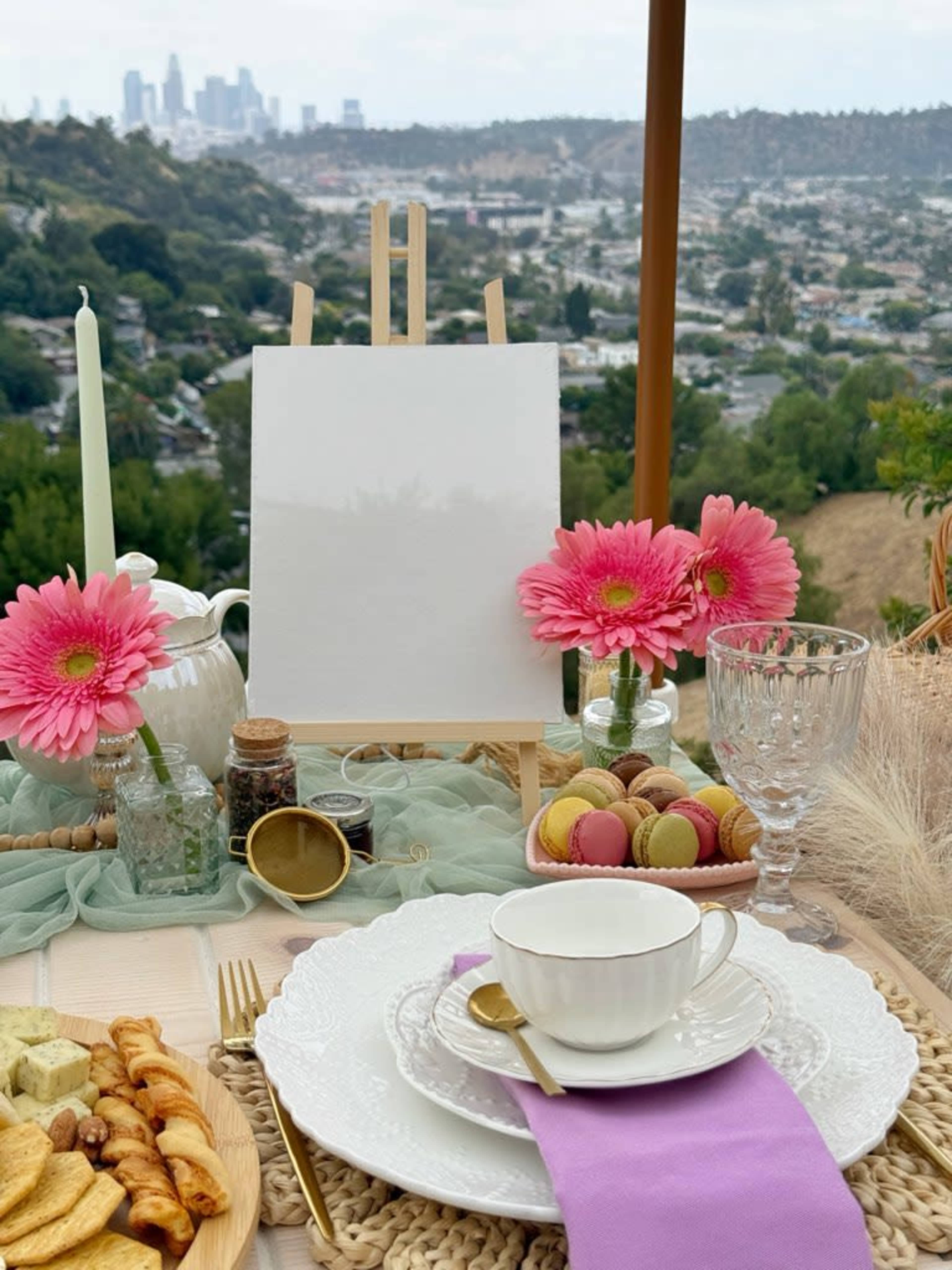 A table is set outdoors with a blank easel, decorative flowers, macarons, and a tea cup against a backdrop of a city skyline in the distance.