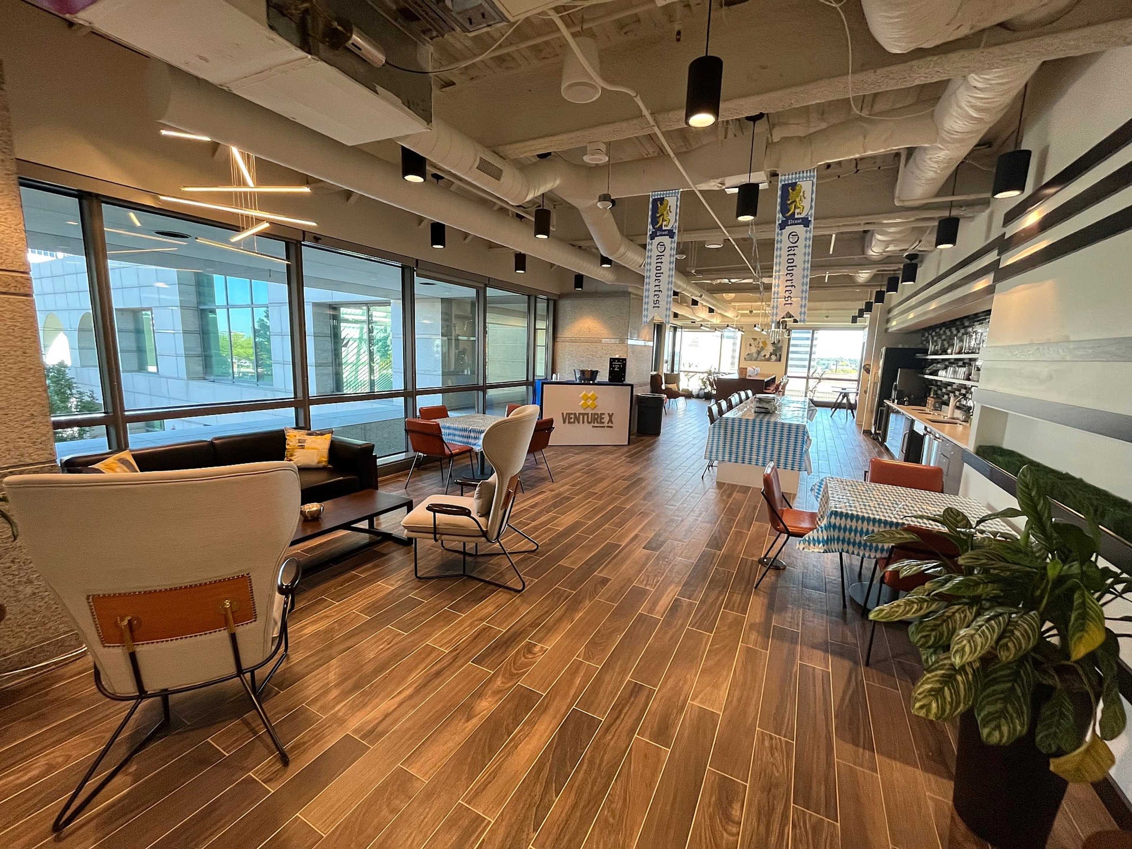 The image shows a modern office lounge area featuring various seating arrangements, large windows, and a decorative plant.