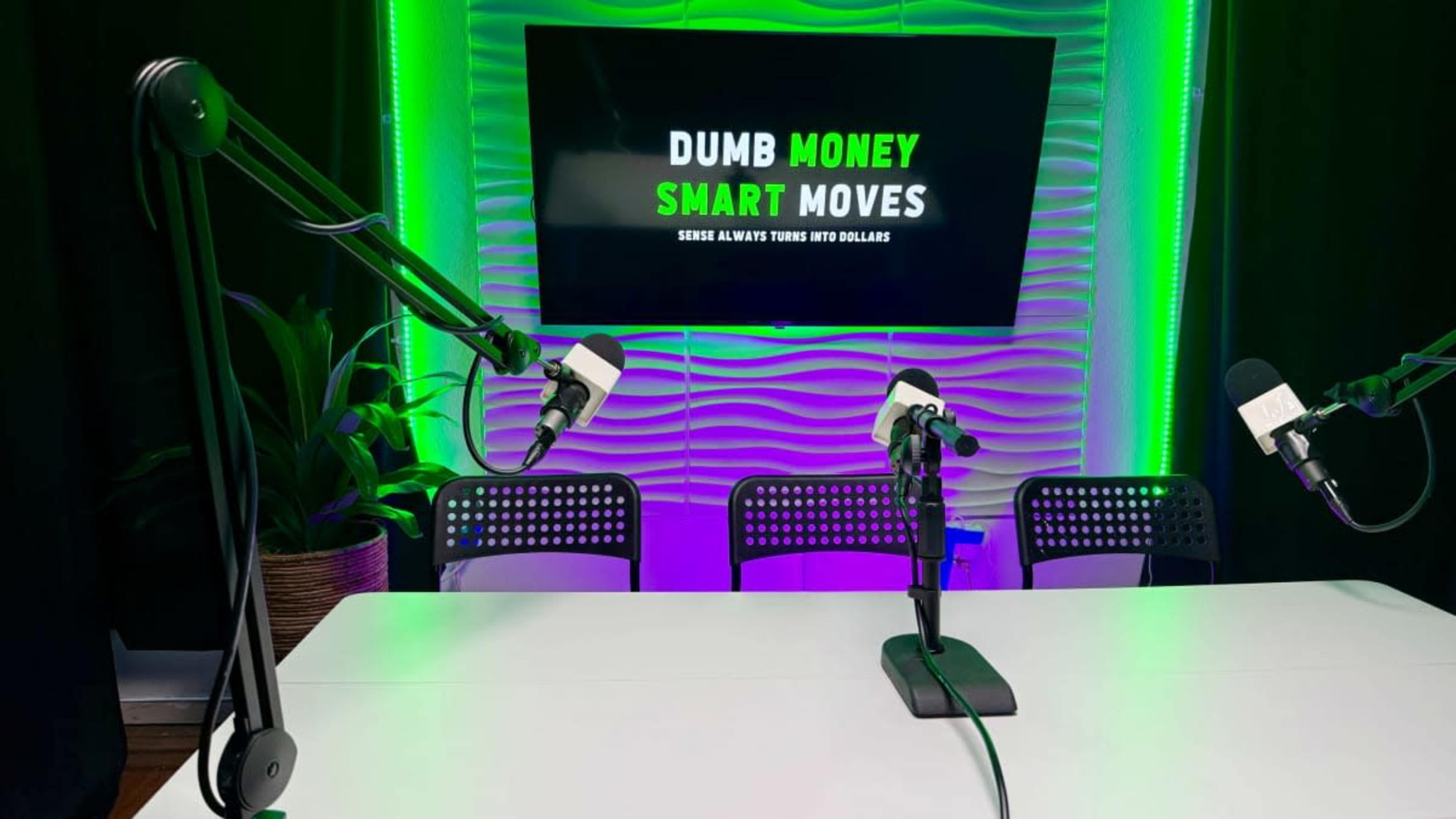 The image shows a podcast recording setup with a table, two microphones, and a backdrop displaying the words "DUMB MONEY SMART MOVES."