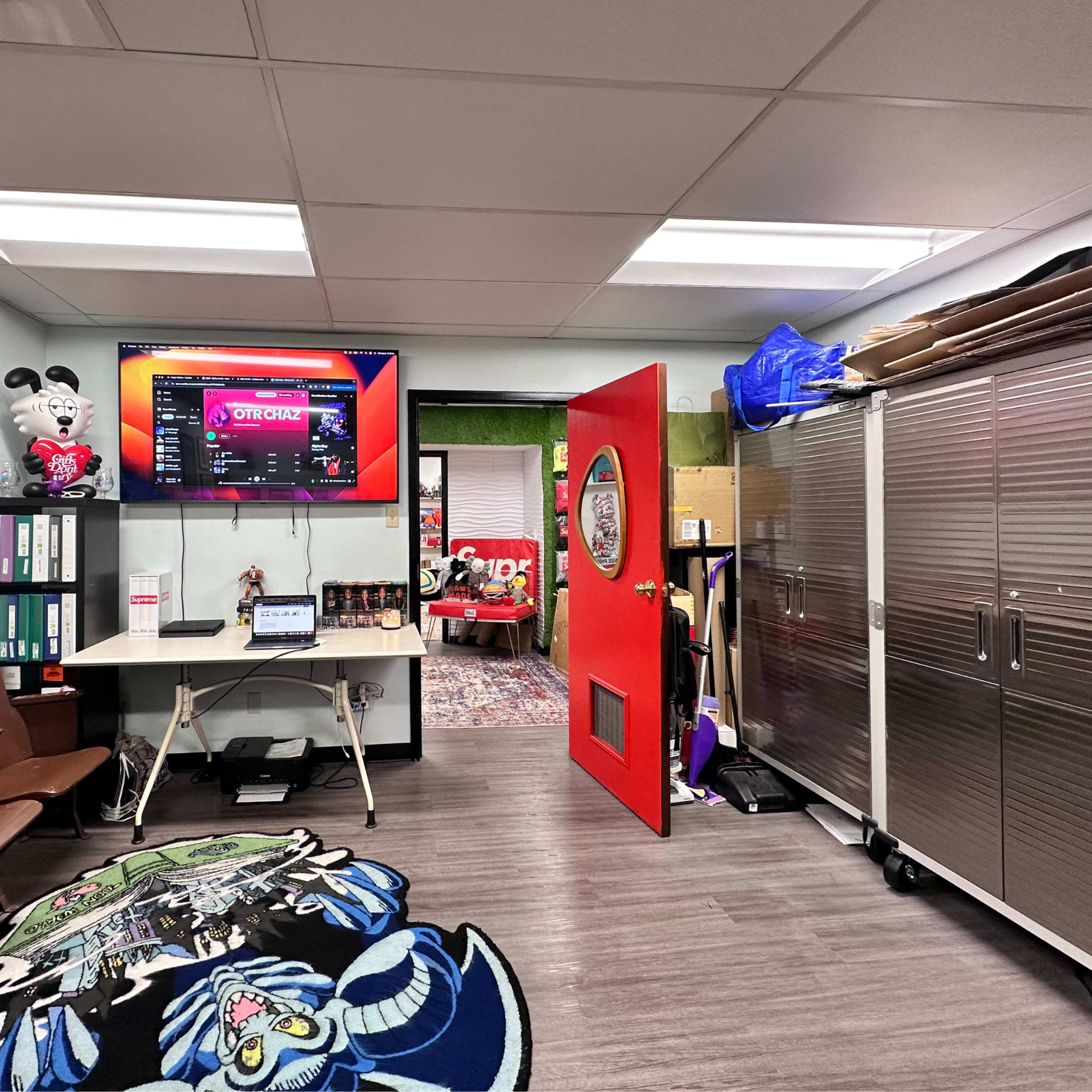 The image shows a brightly colored office space with a red door, a large screen displaying a Spotify playlist, and metal storage cabinets along one wall.
