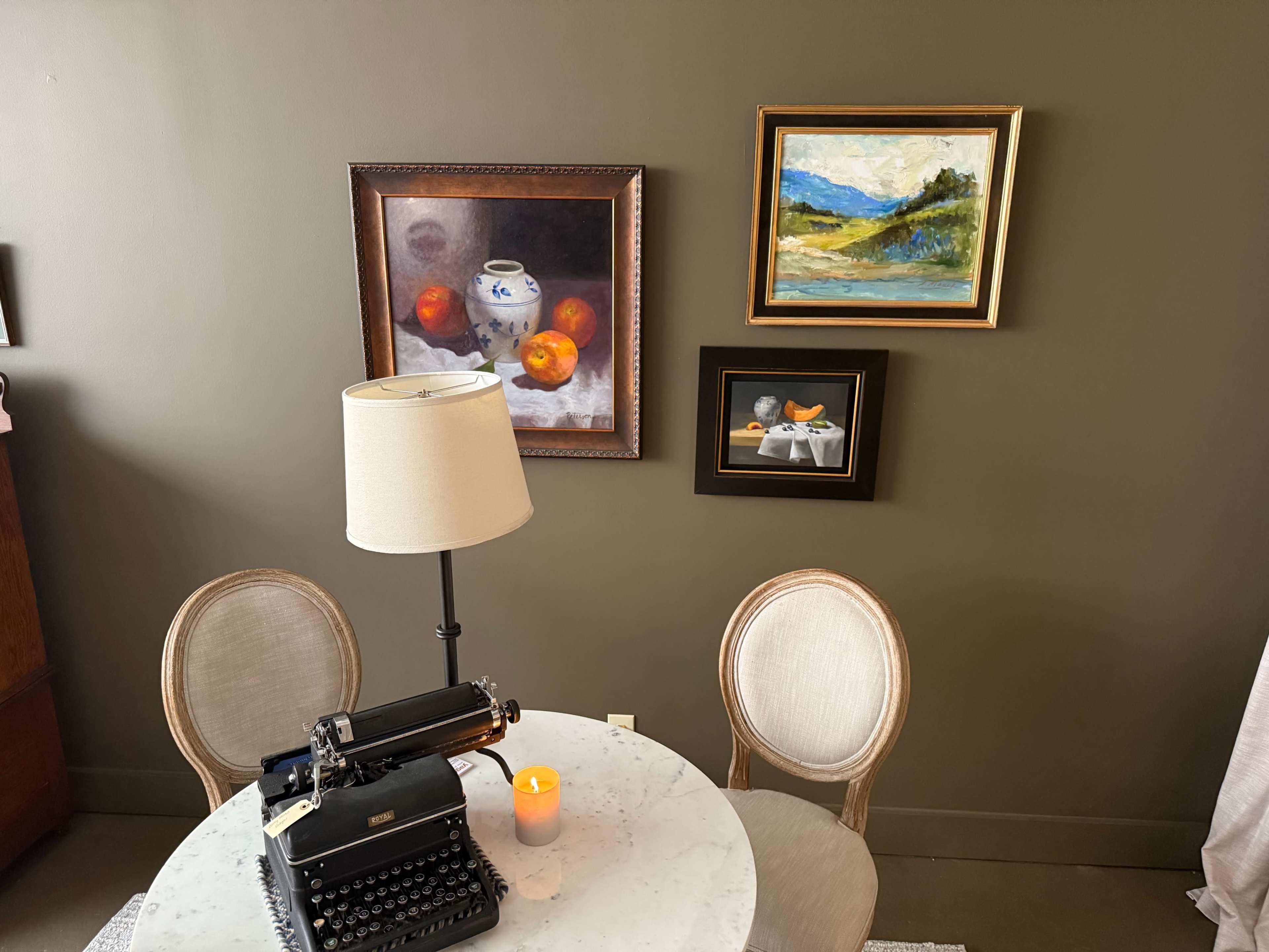 A vintage typewriter sits on a circular table beneath a lamp, surrounded by framed artwork and two upholstered chairs in a softly lit room.