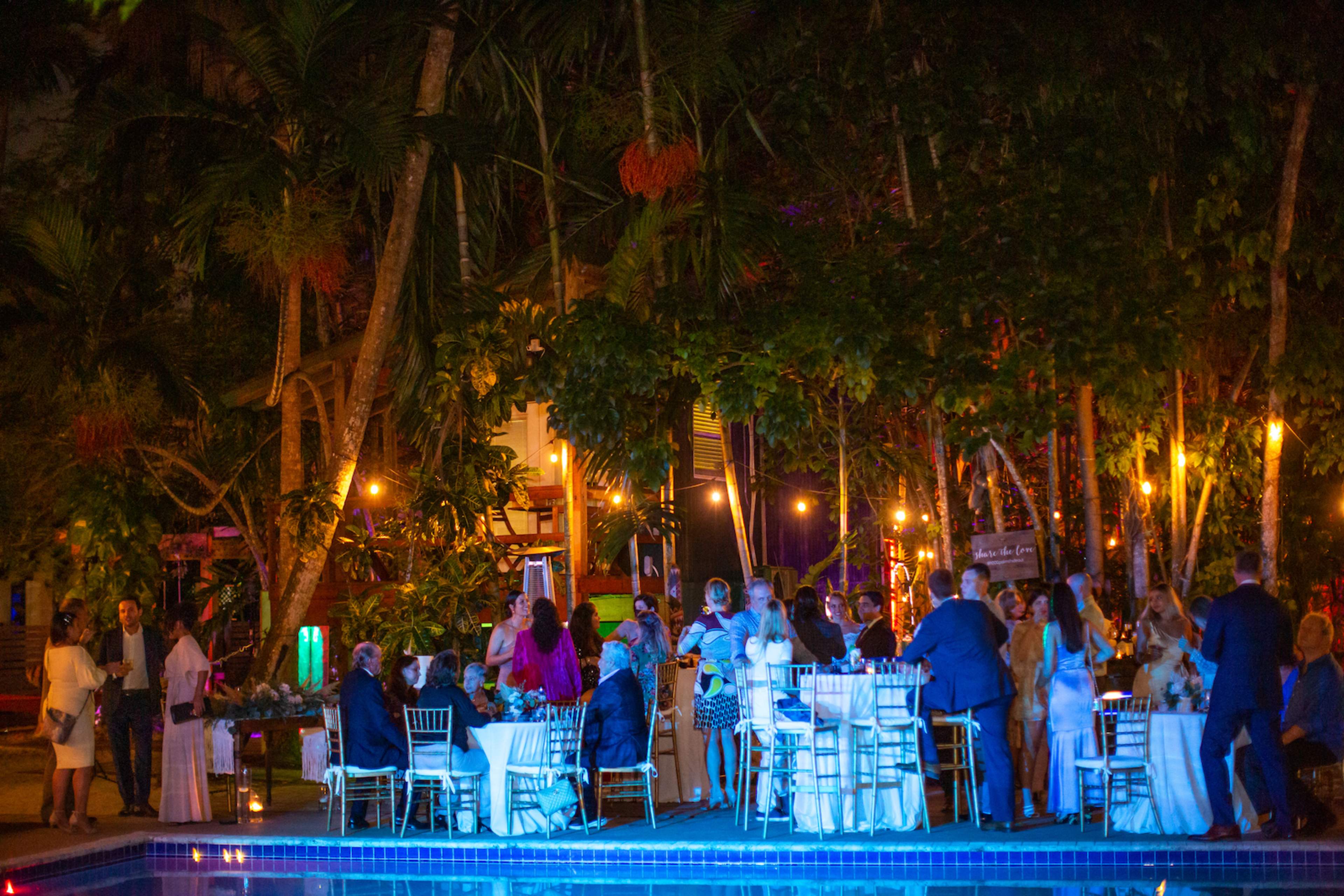 A group of people gathers around tables by a pool in a tropical setting at night, illuminated by colorful lights.