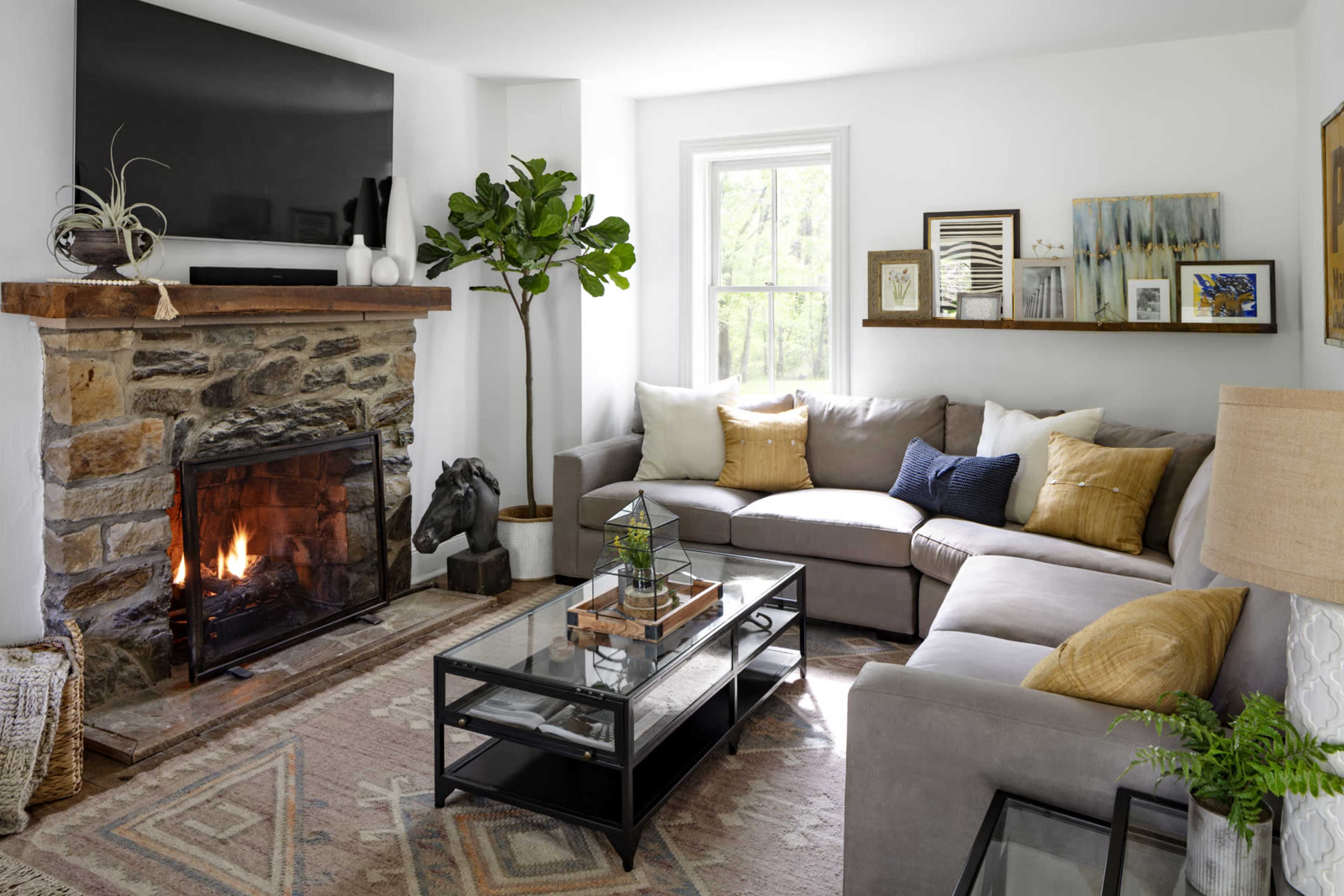 The living room features a stone fireplace with a roaring fire, a sectional sofa, a glass coffee table, and decorative plants.
