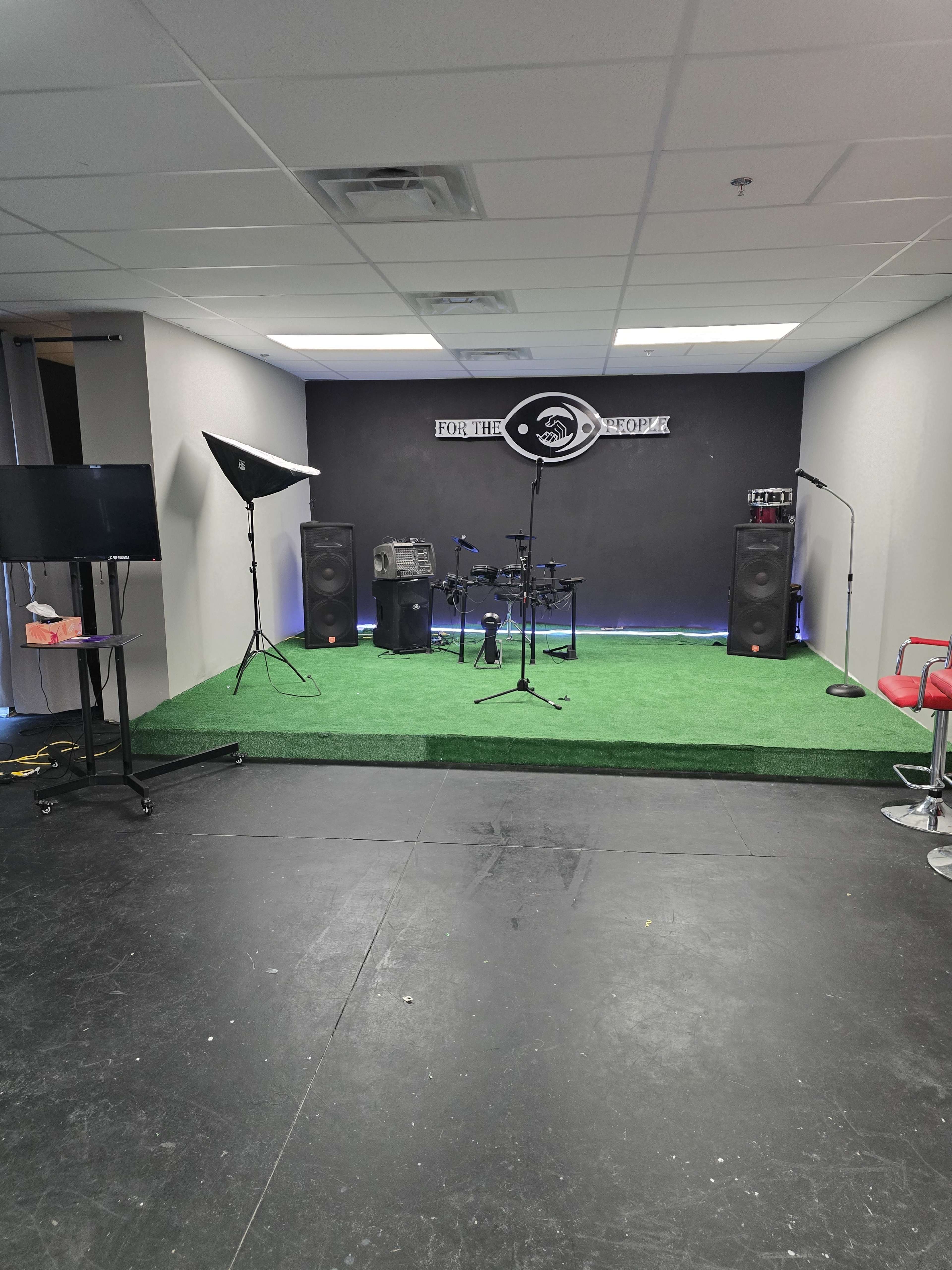 A small stage area with musical equipment, including a drum set and speakers, set against a black wall and green carpeting.