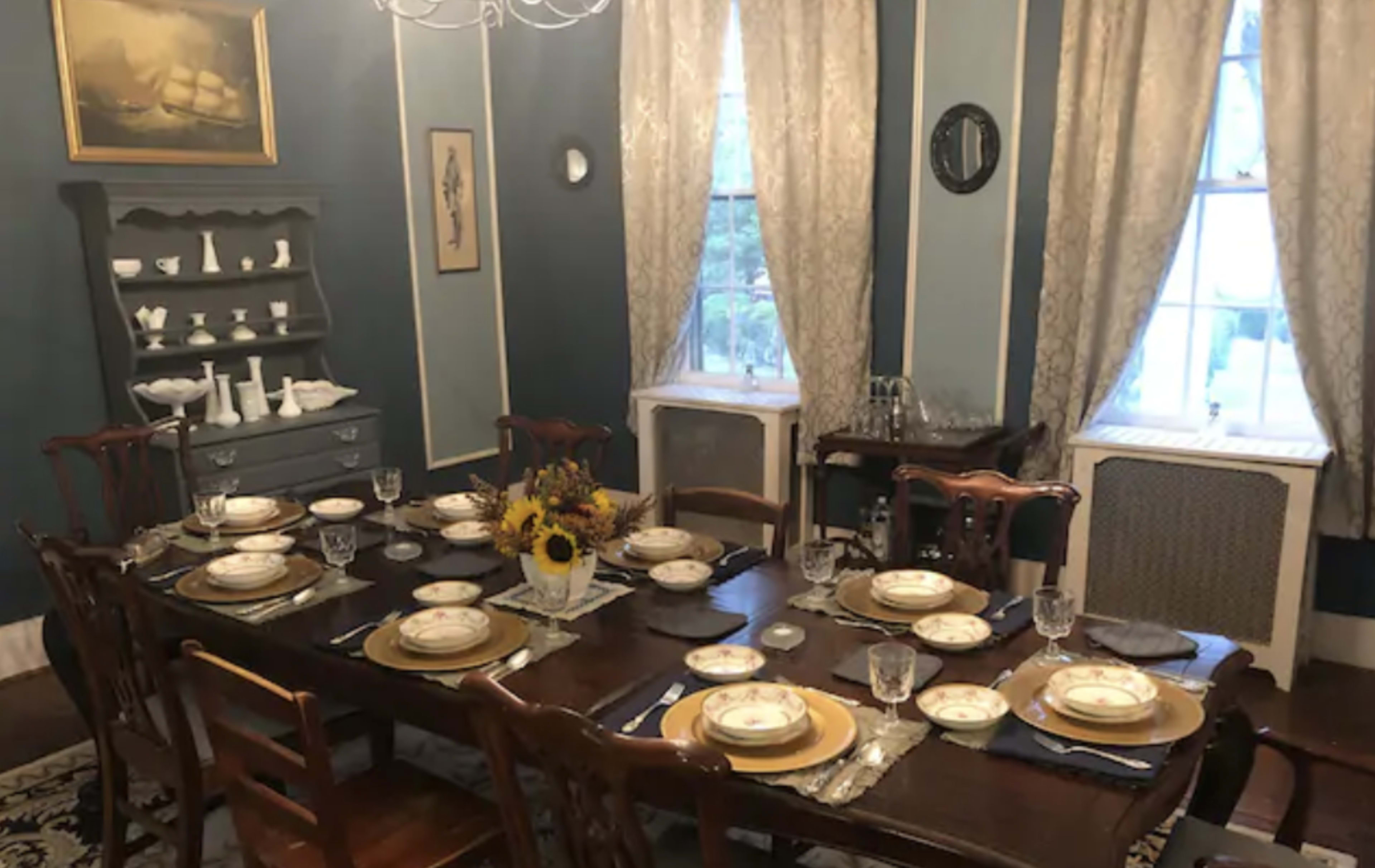 A formal dining room is set with a large wooden table surrounded by chairs, adorned with plates, glasses, and a vase of flowers, while blue and white walls frame the scene.
