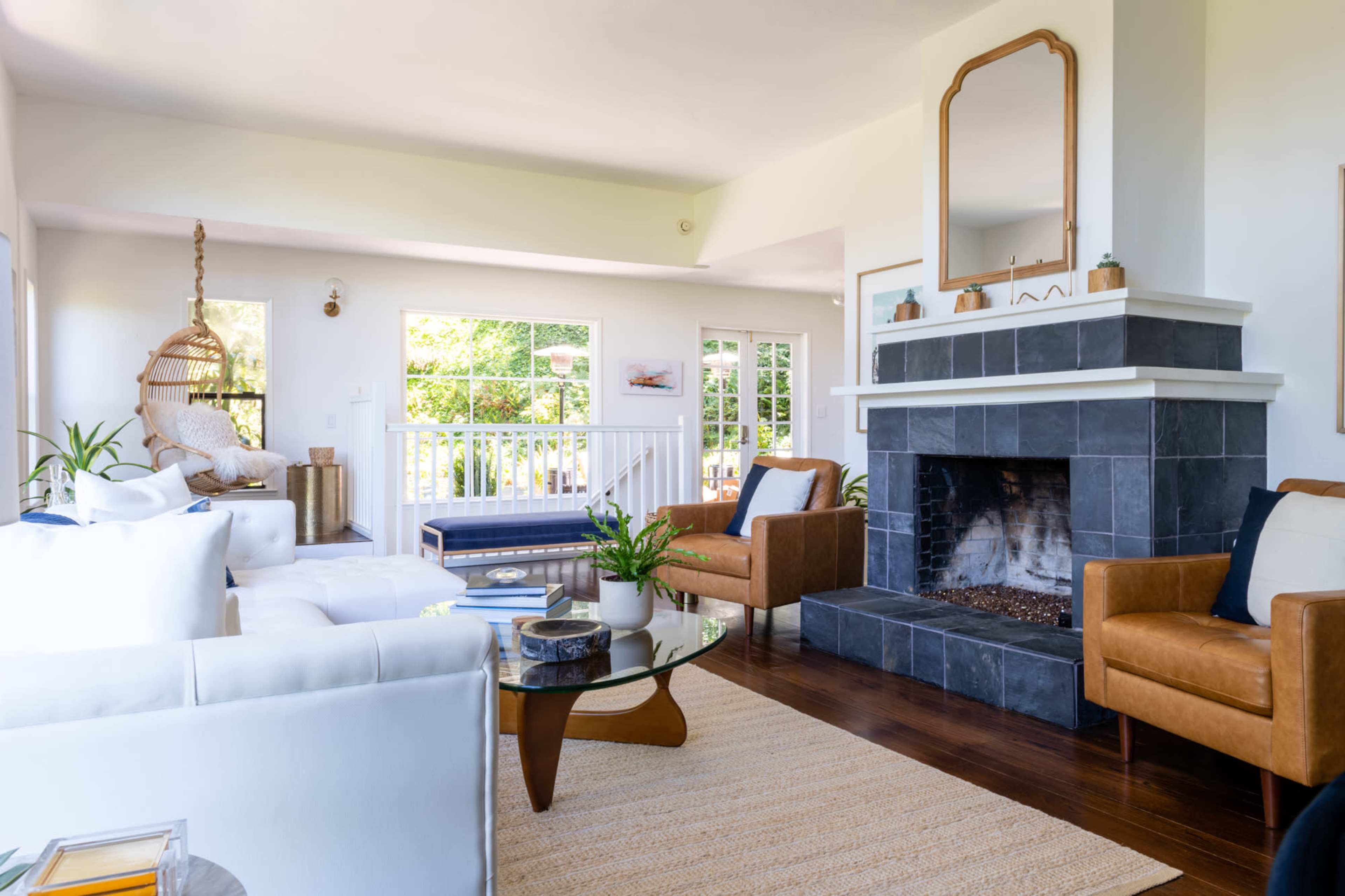 The image shows a modern living room with a fireplace, white sofas, leather armchairs, and large windows allowing natural light.