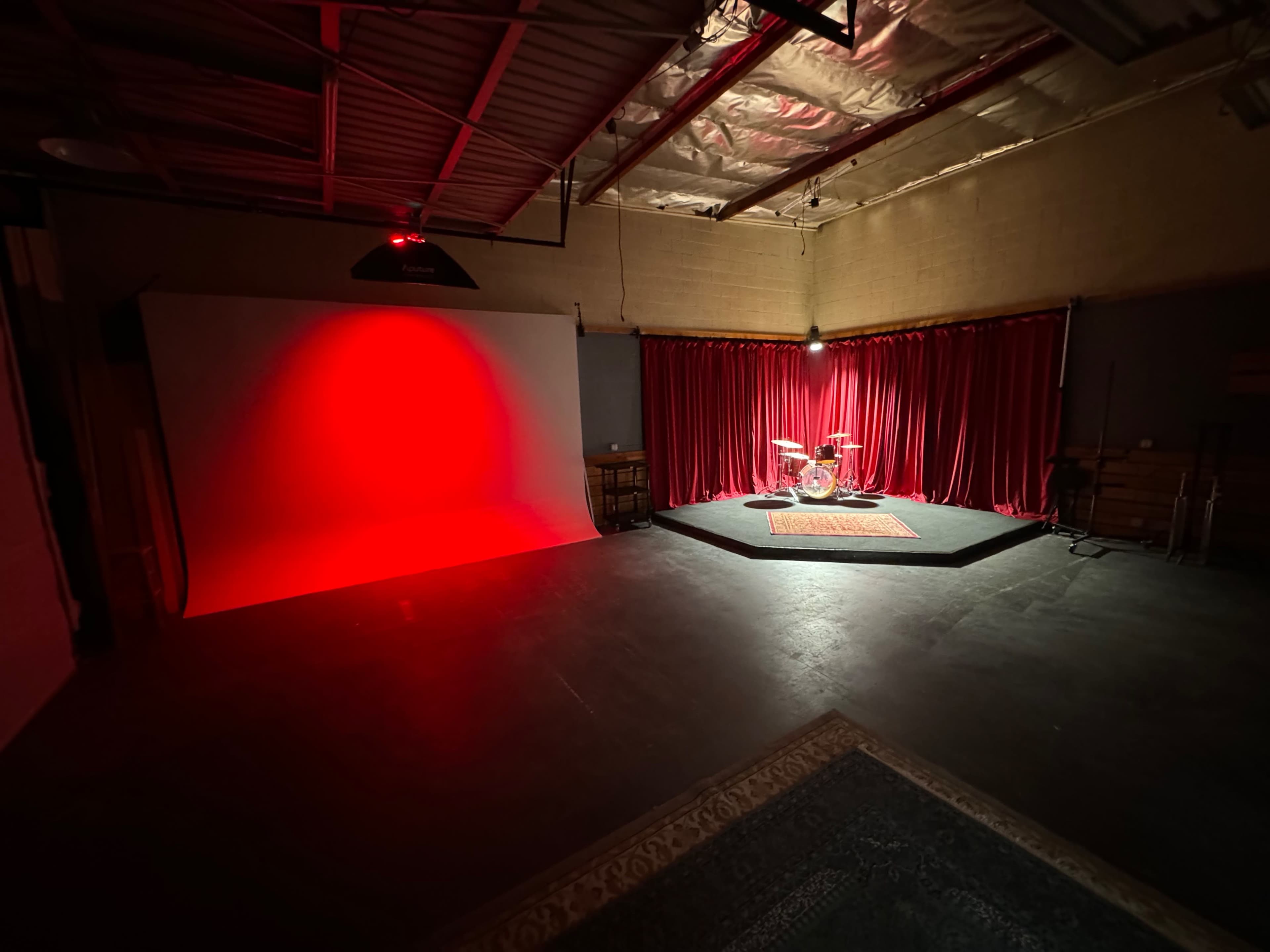 The image features a spacious studio with a red-lit backdrop, red curtains, and a drum set positioned on a raised platform.