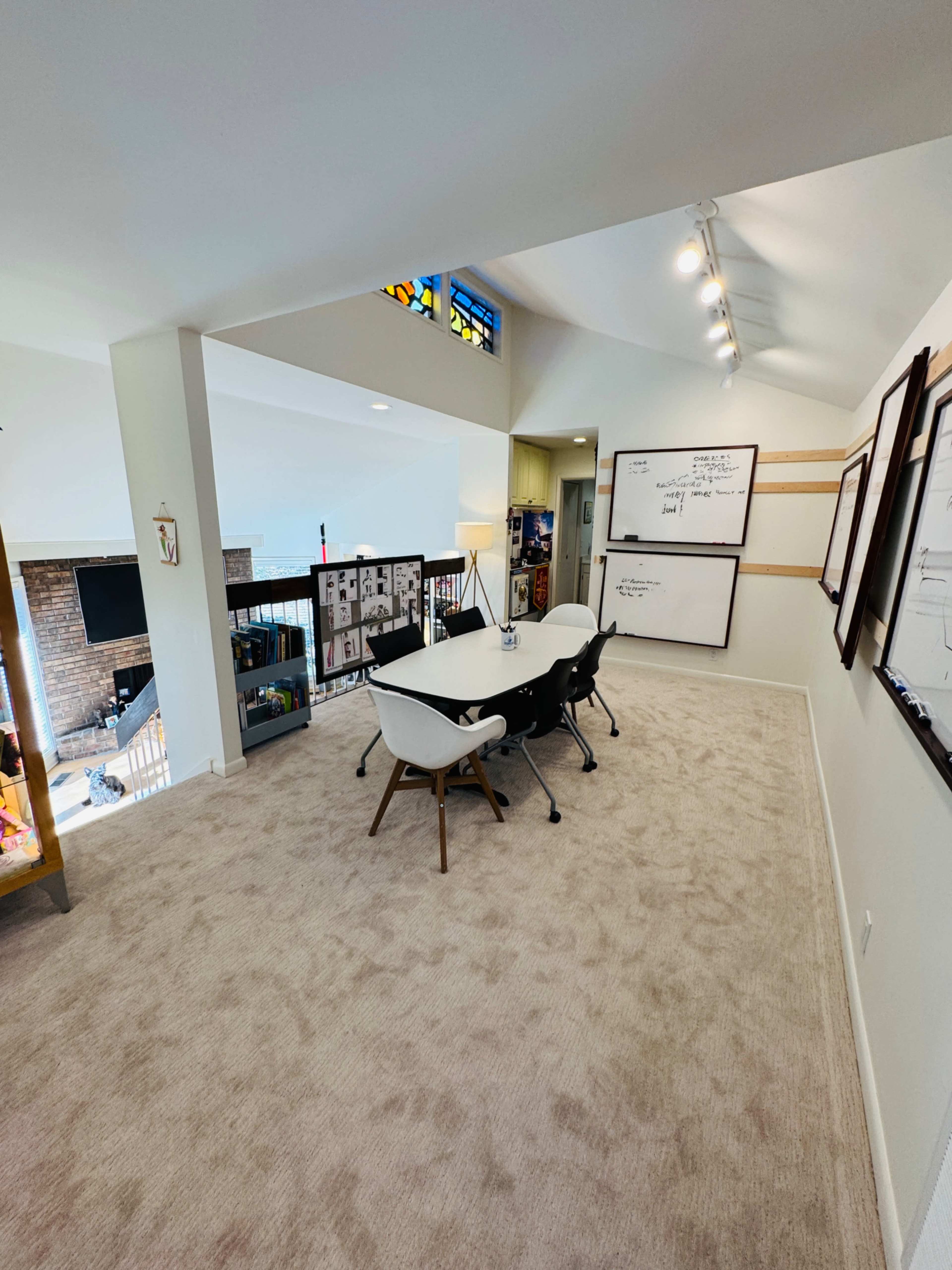 A spacious meeting area is furnished with a white table and black chairs, featuring whiteboards on the walls and a stained glass window above.