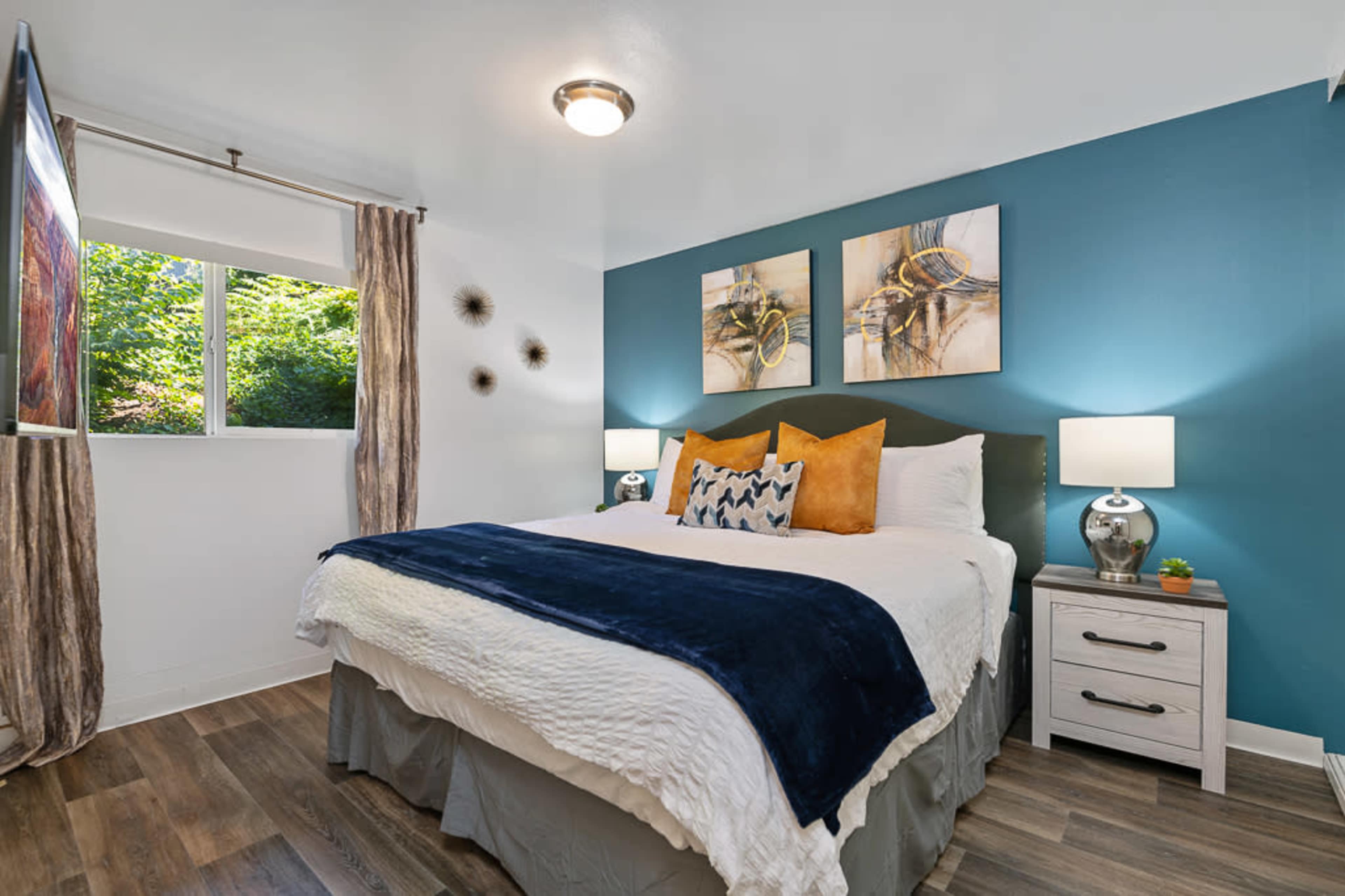 The bedroom features a bed with a white quilt, blue and orange accents, and framed artwork on the wall, alongside a window and bedside tables with lamps.