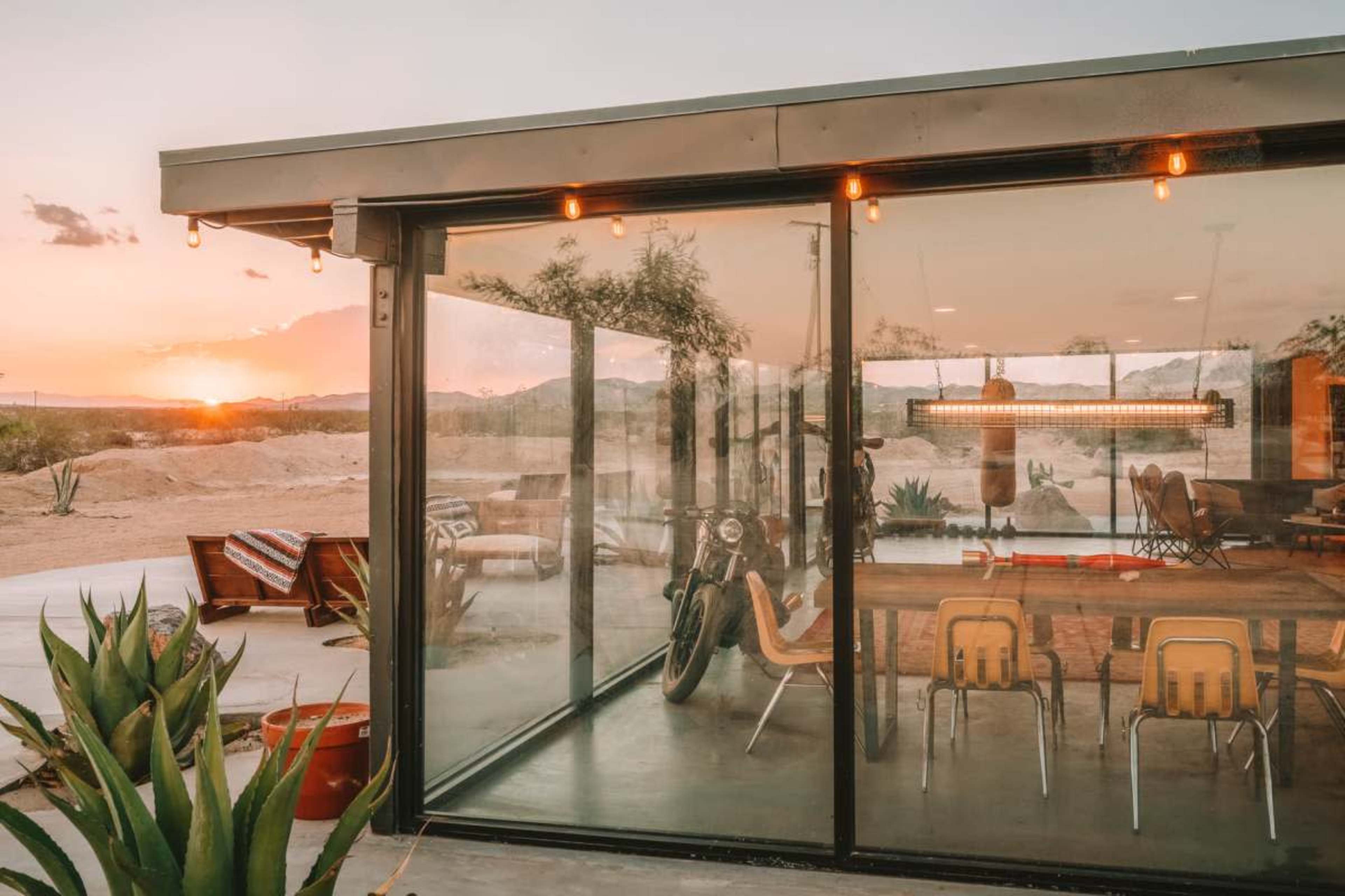A modern glass-walled building showcases an interior dining area with a motorcycle, framed by a desert sunset.