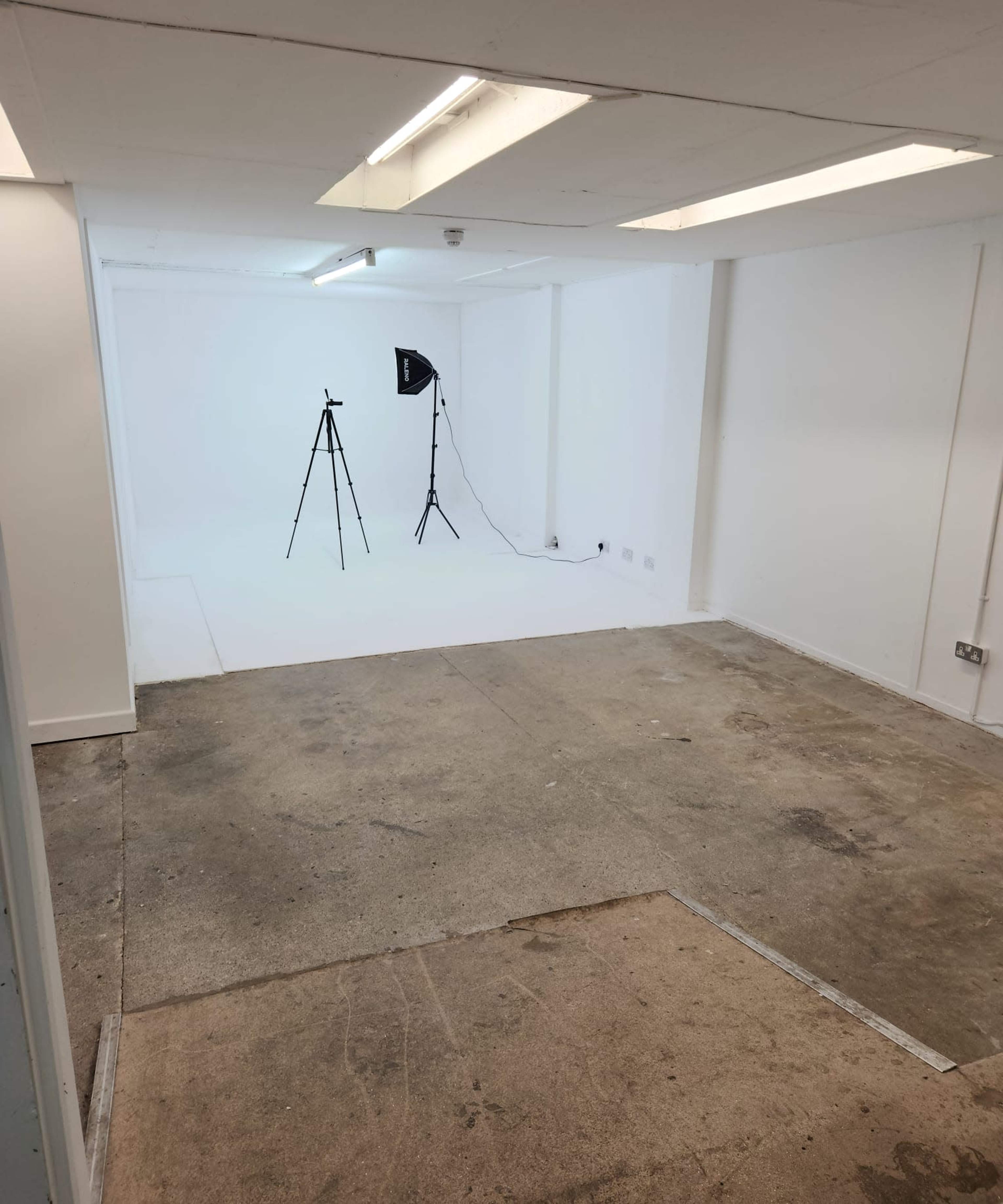 The image shows a sparse, empty studio space with a concrete floor and white walls, featuring two tripods and a light setup in the corner.
