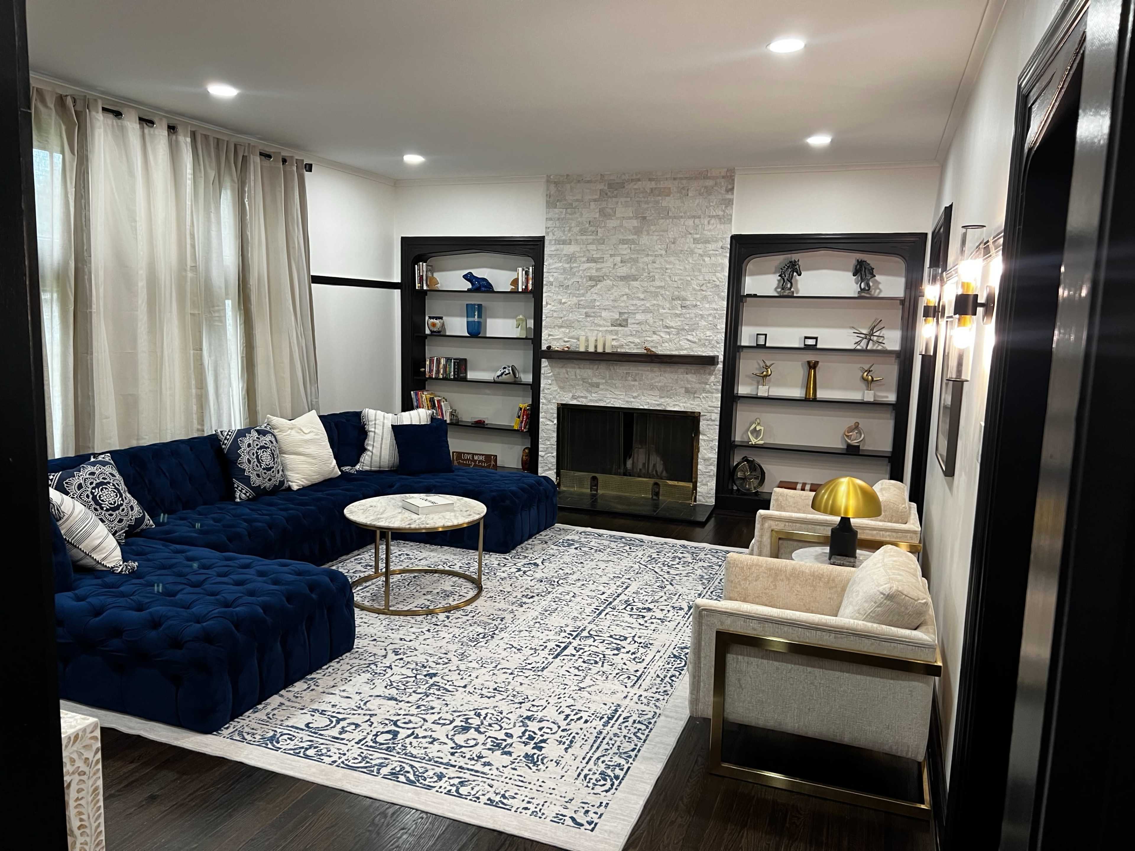 The image shows a modern living room with a blue tufted sectional sofa, a marble coffee table, a large area rug, and a stone fireplace surrounded by built-in shelves.