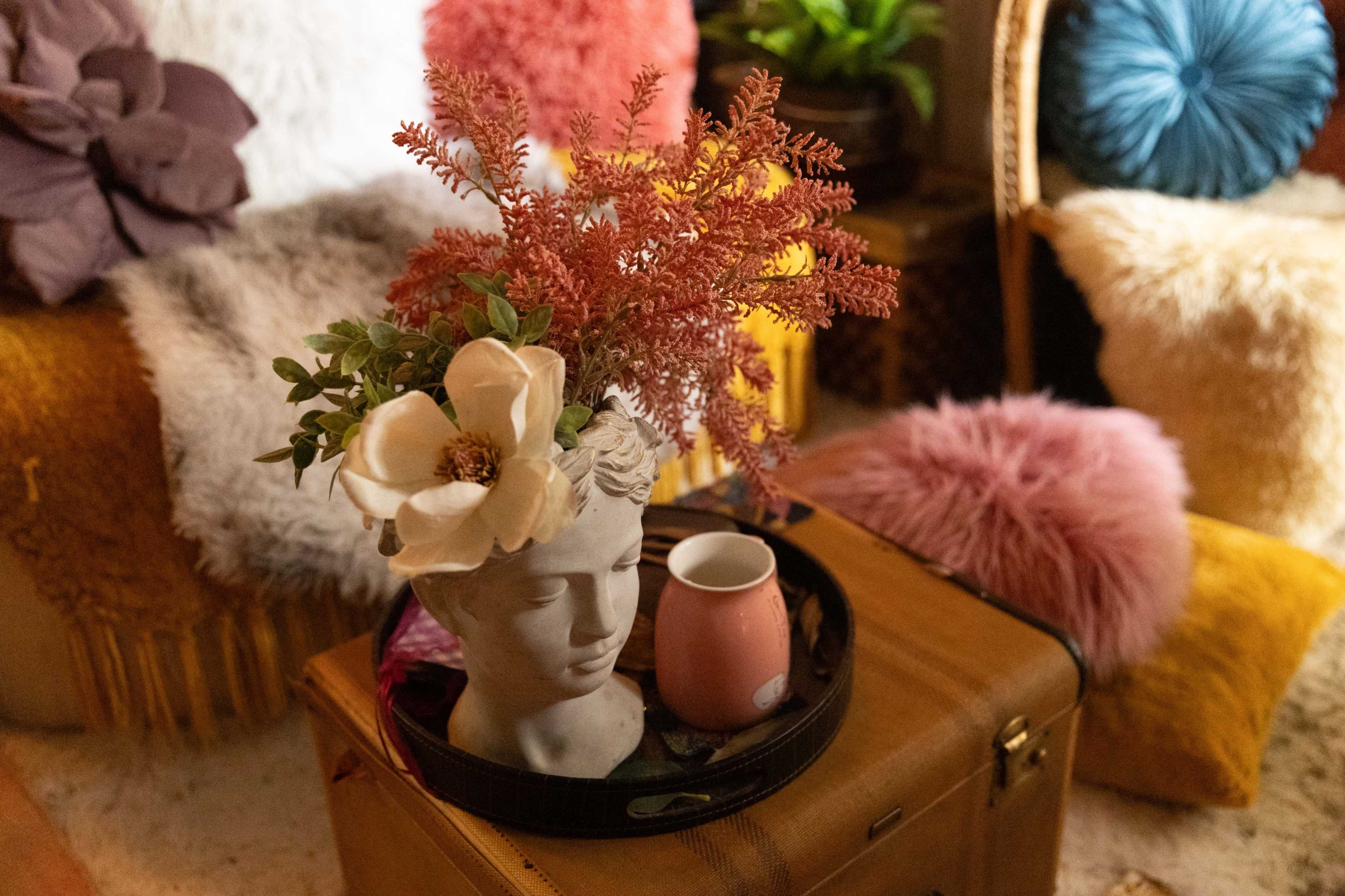 A decorative head vase with flowers sits on a black tray next to a pink ceramic cup, surrounded by plush cushions and warm textured fabrics.