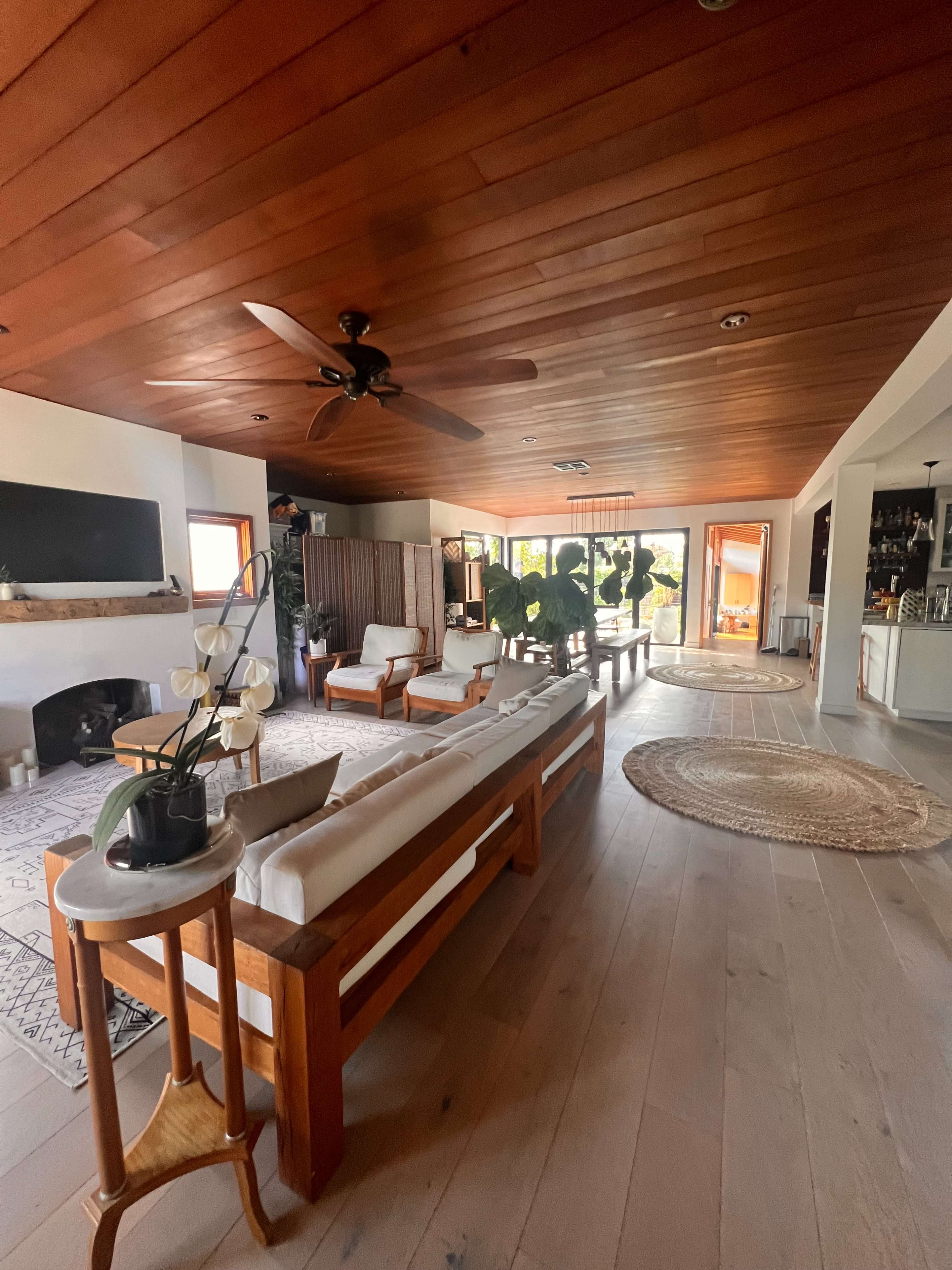 The spacious living room features a wooden ceiling, a large window with sliding doors, and various seating arrangements including a sectional sofa and armchairs.
