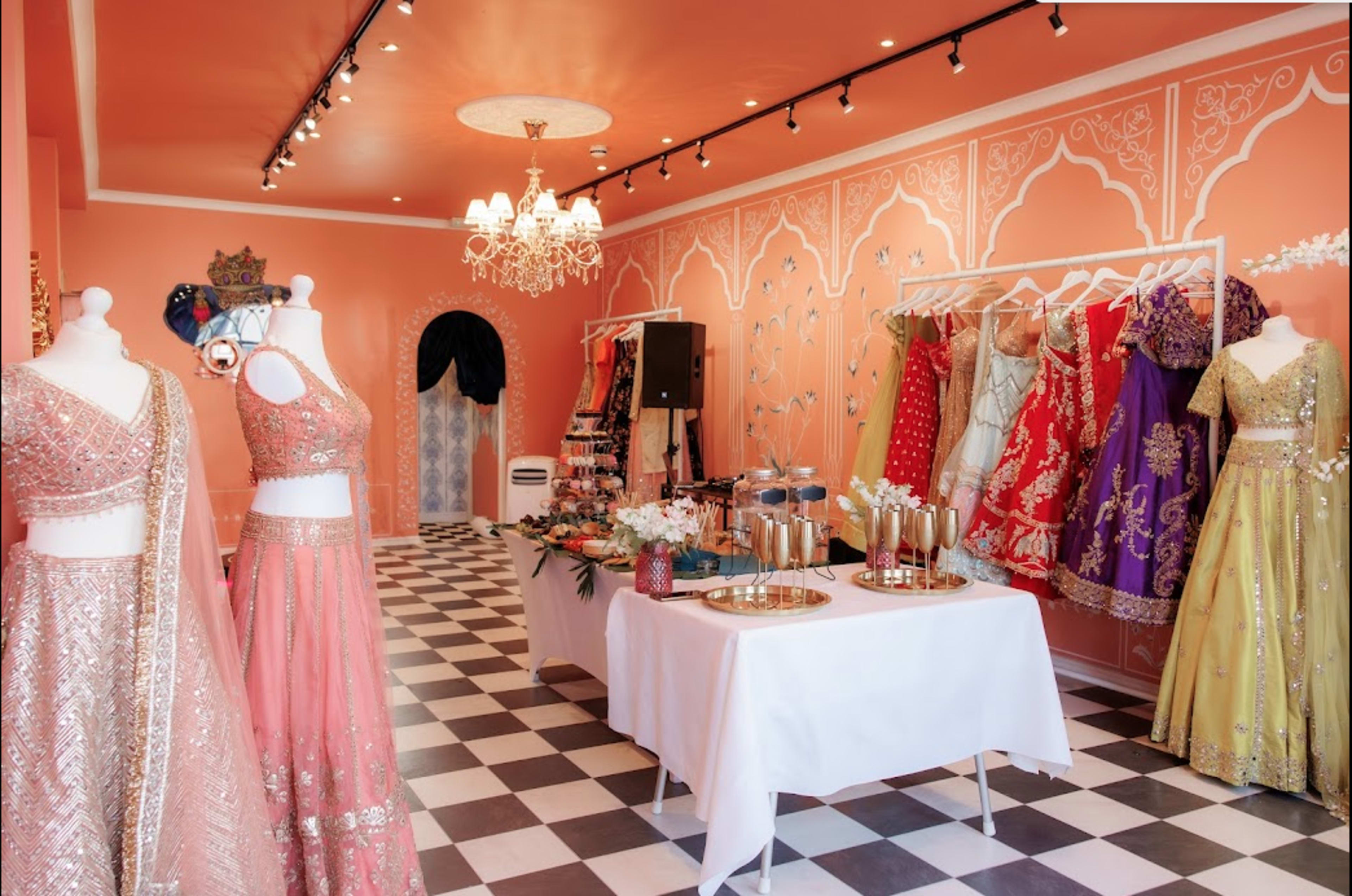 A boutique interior featuring elegant traditional dresses displayed on mannequins and racks, with a checkered floor and decorative wall patterns in soft coral tones.