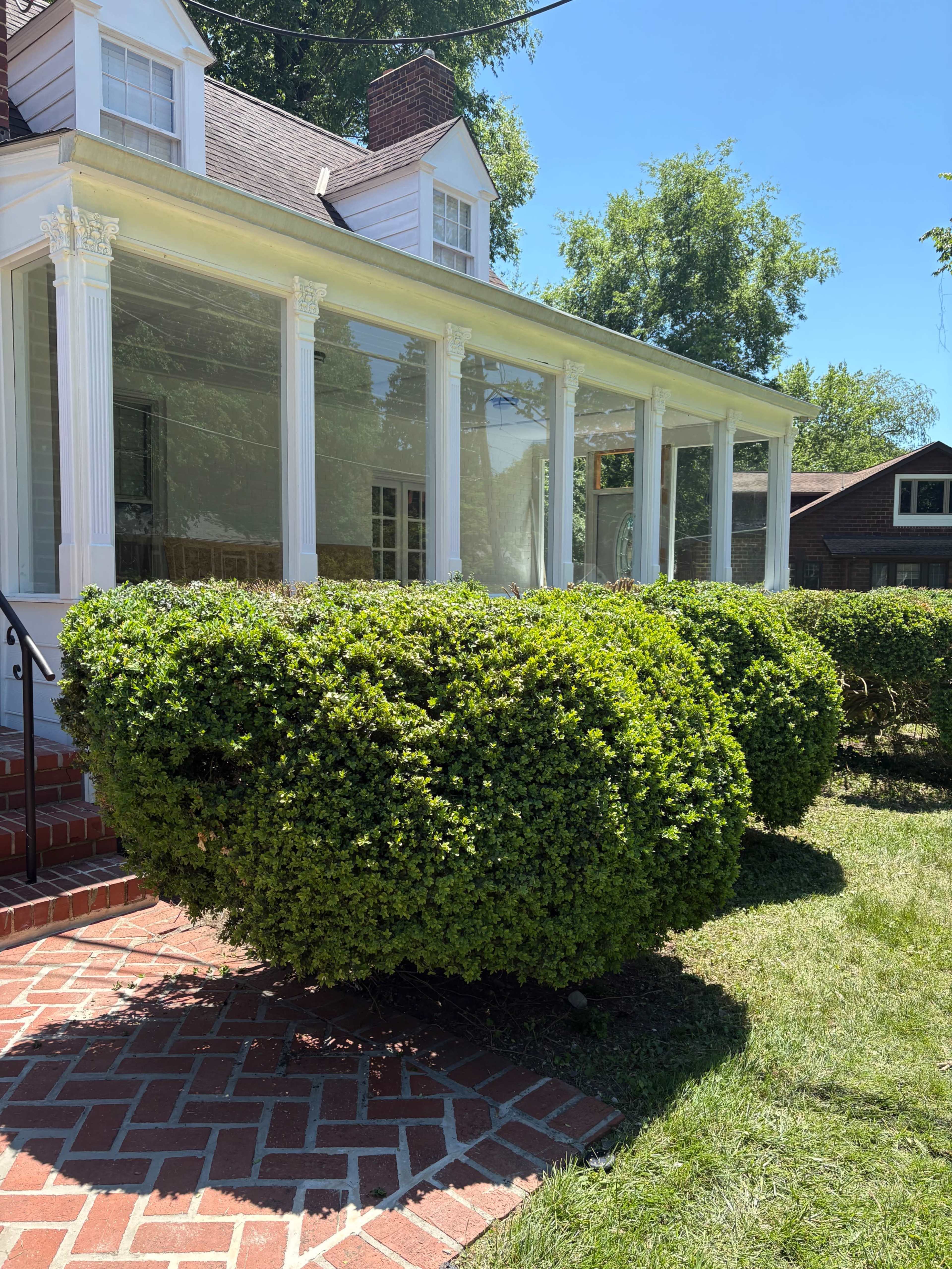 A well-maintained house with a porch features neatly trimmed, rounded bushes in the foreground and a lawn with grass.