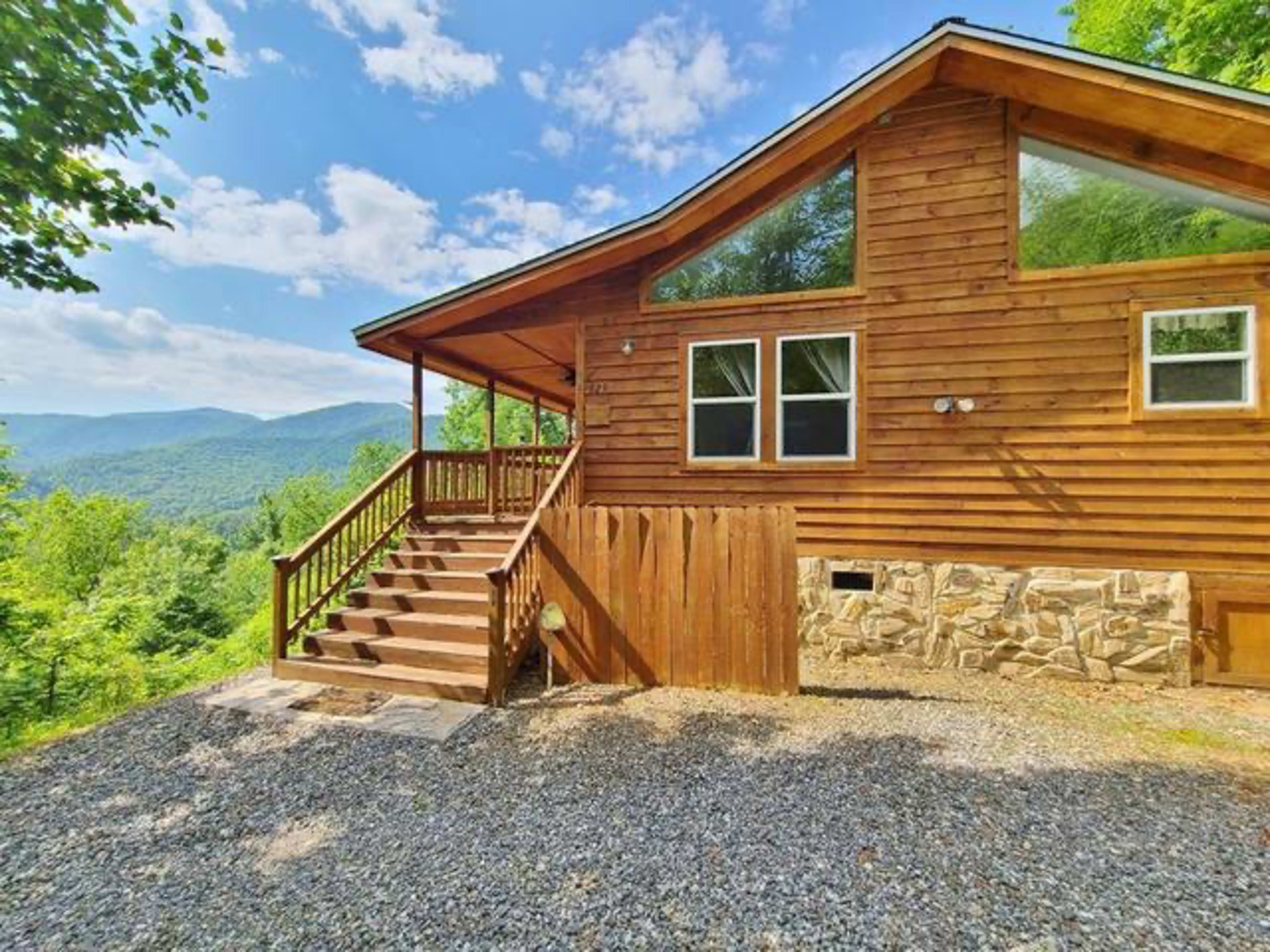 A wooden cabin with a stone foundation and large windows overlooks a mountainous landscape under a clear blue sky.