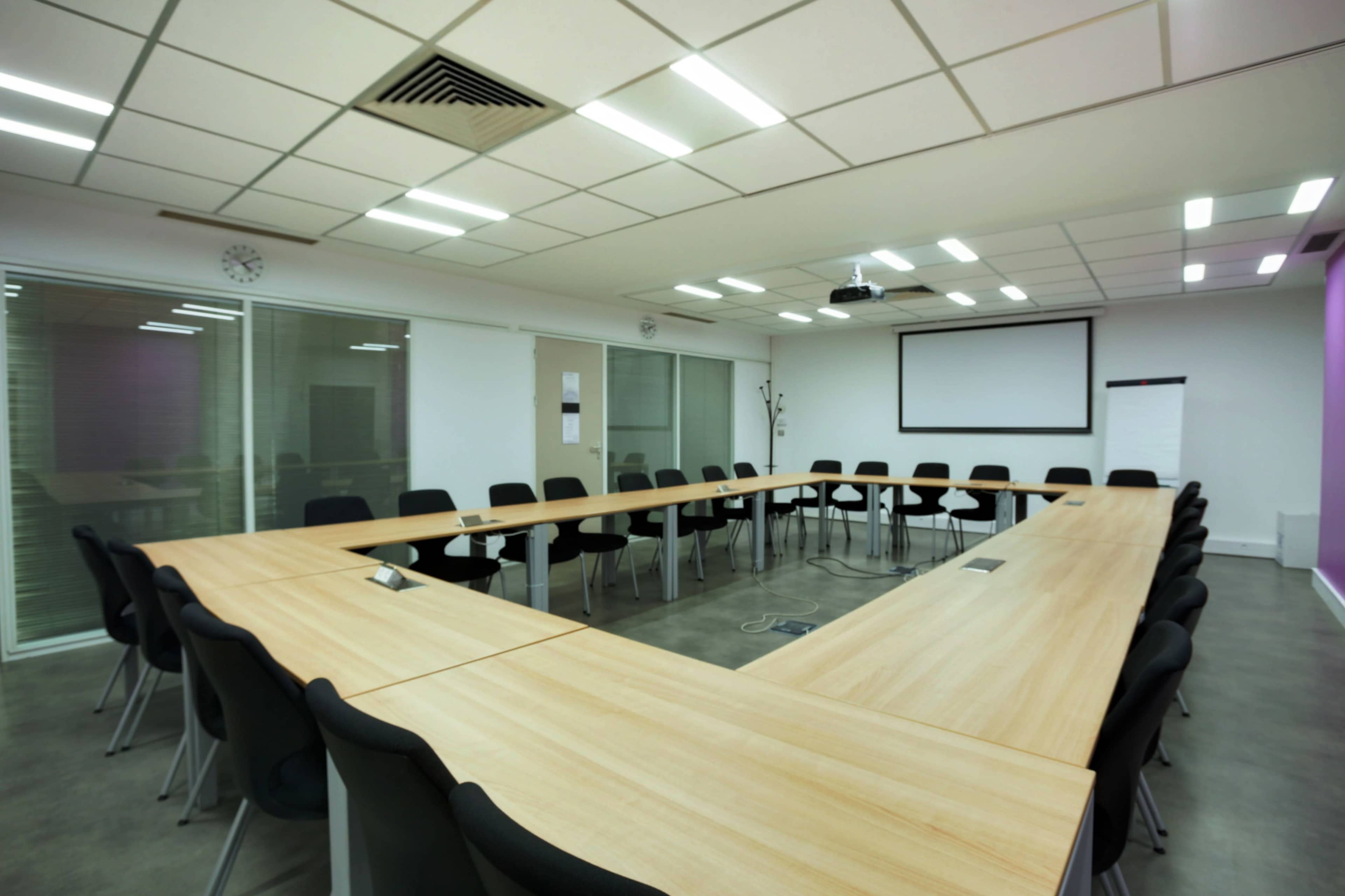 A large conference room features a U-shaped arrangement of wooden tables and black chairs, with a projector screen at one end and glass windows along the walls.