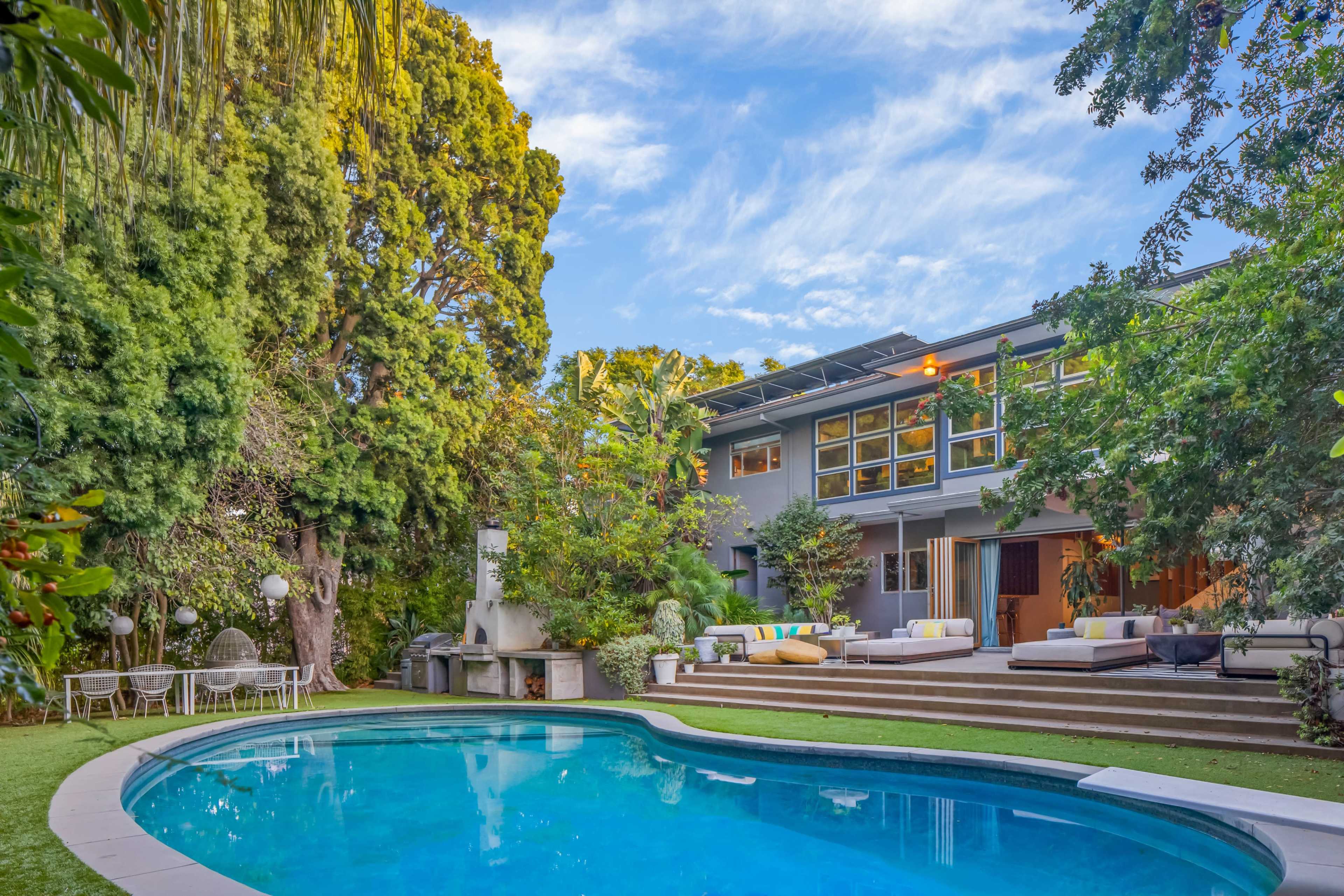 The image shows a modern house with large windows surrounded by lush greenery and a curved swimming pool in the foreground.