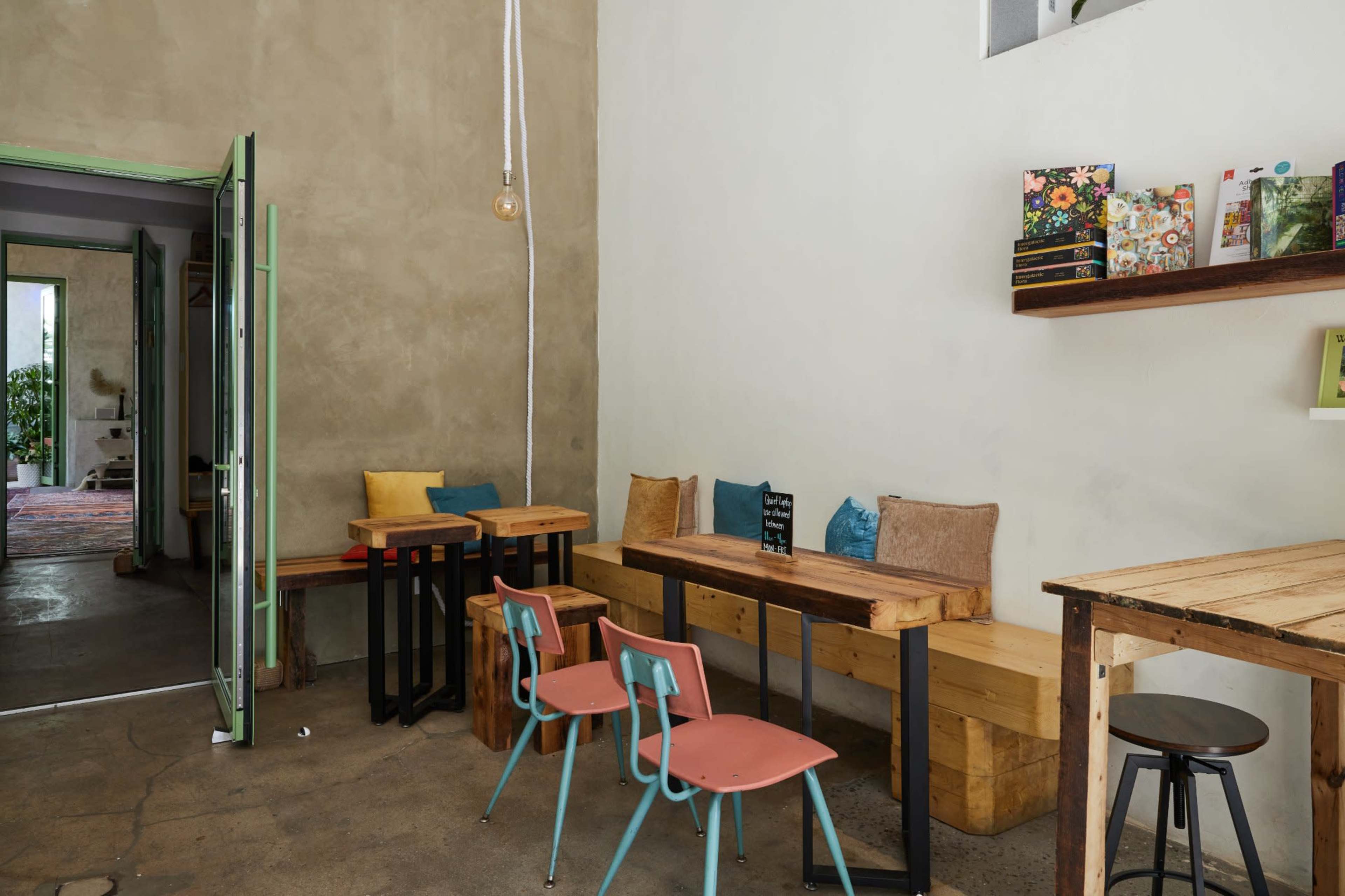 The image shows a minimalist café interior with wooden tables, colorful chairs, and a simple, light-colored wall.