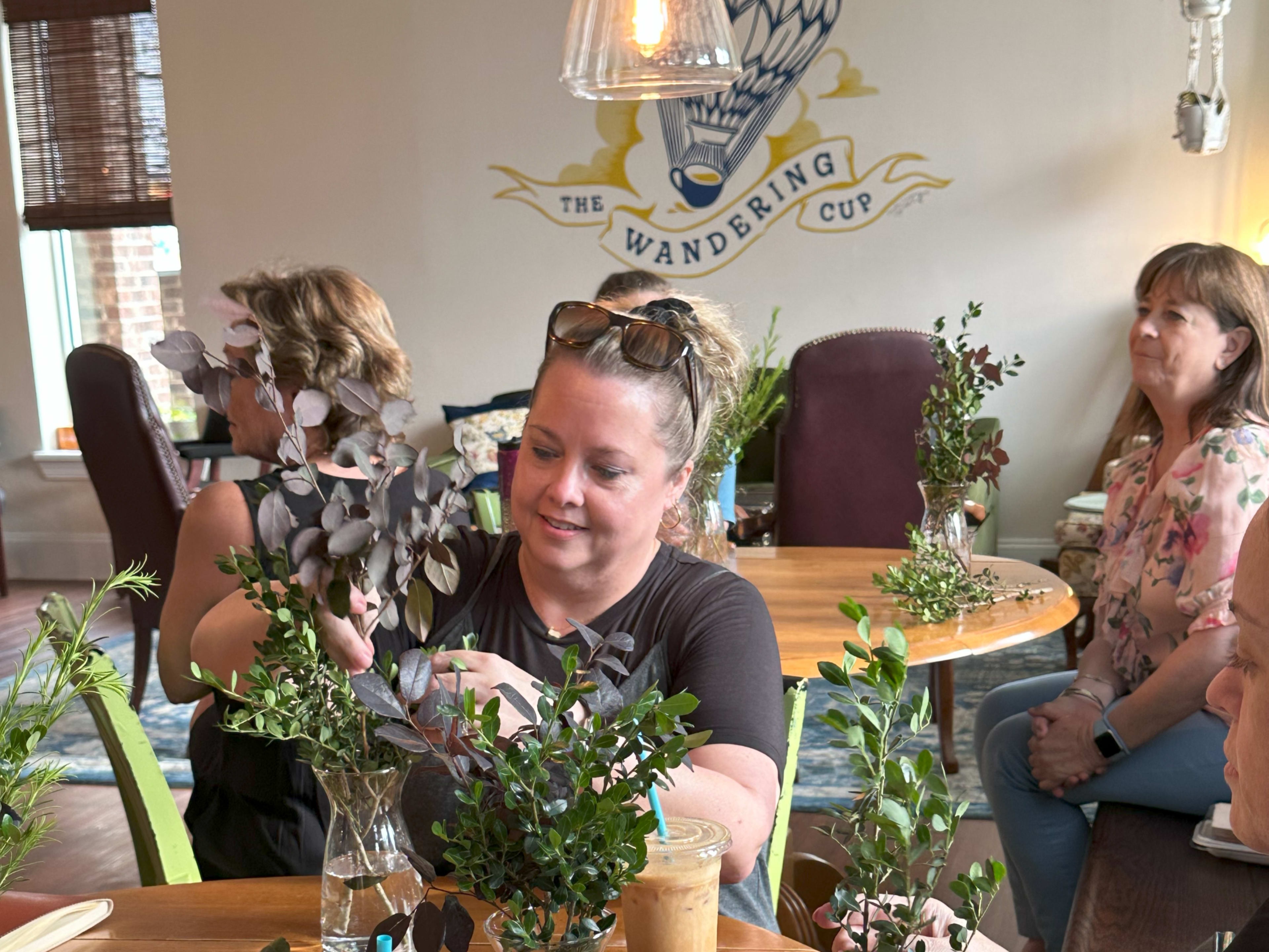 A woman arranges greenery in a glass vase at a table while others observe in a café decorated with a mural.