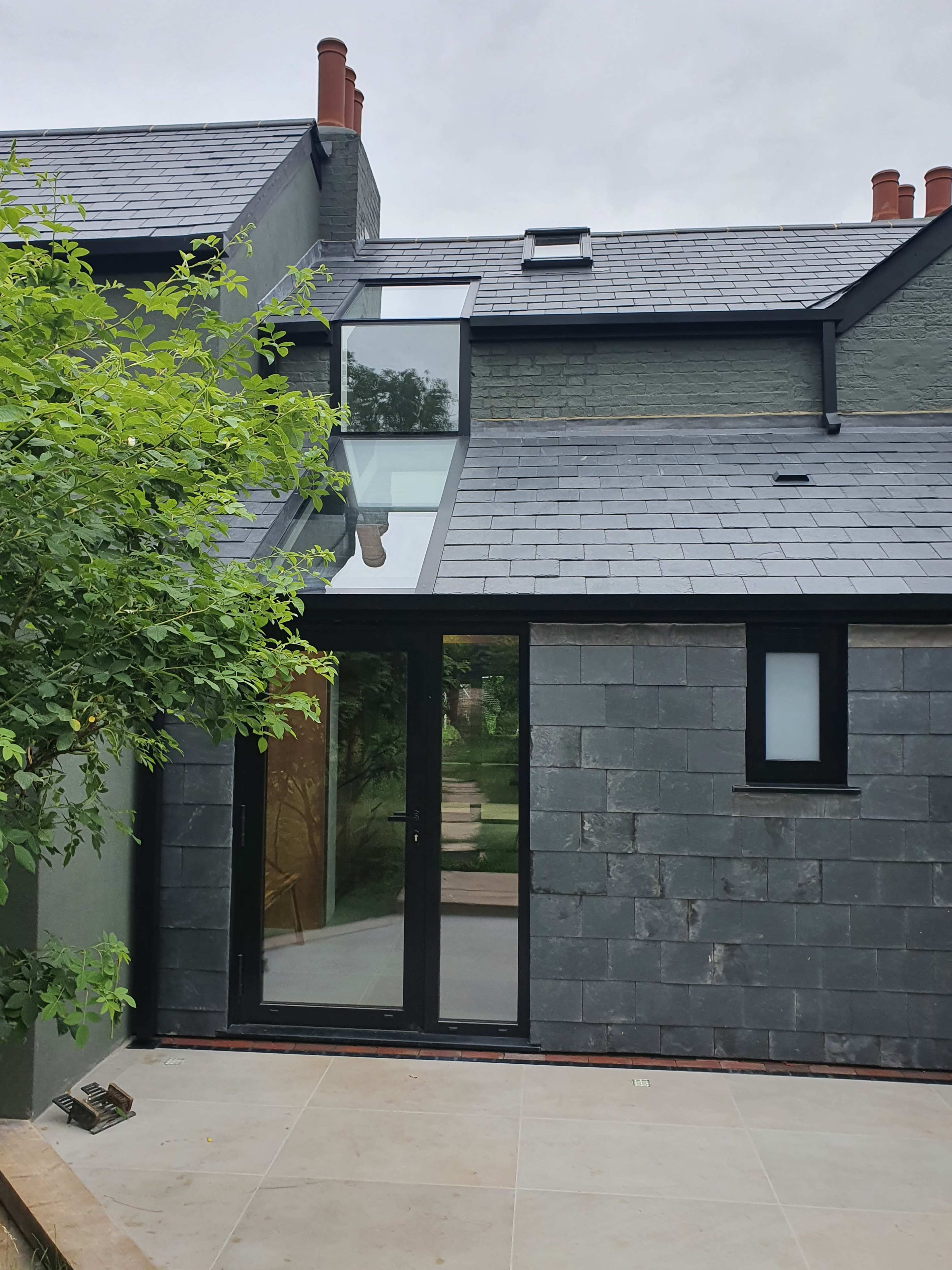 A modern house exterior with a slate roof, large glass doors, and a tree partially obscuring the view.