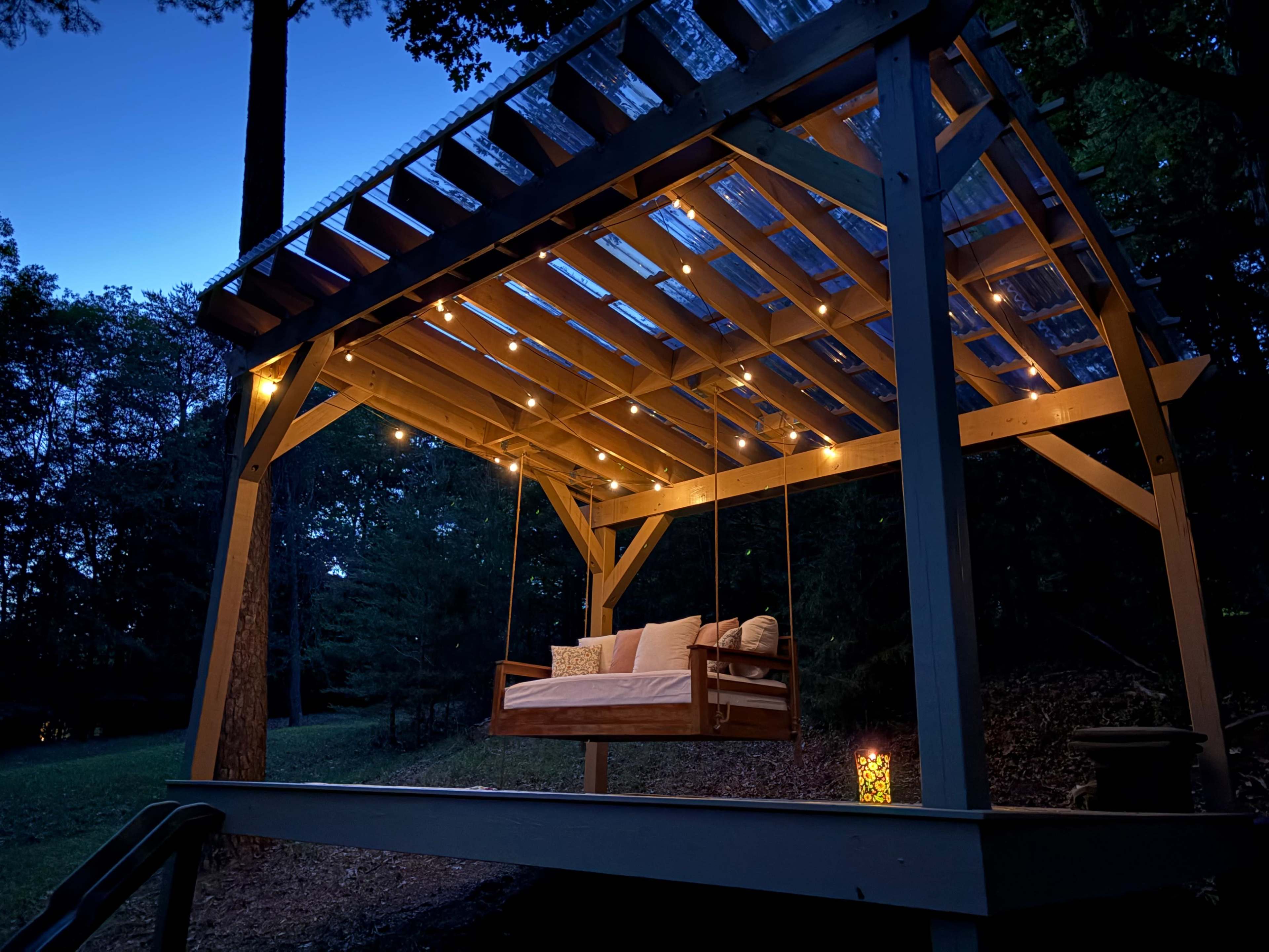 A wooden swing bed with cushions is illuminated by string lights under a slatted roof in a wooded area during twilight.